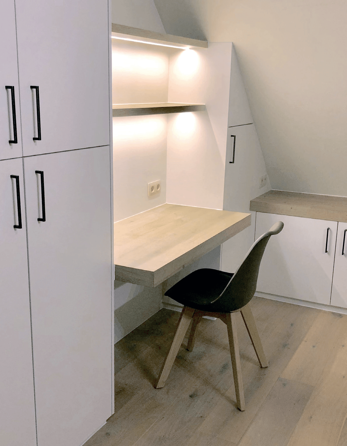 Wit ingebouwd bureau met kasten en een zwarte bureaustoel. Houten bureaublad, zachte verlichting.