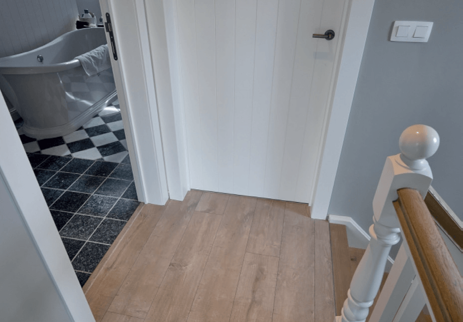 VDH Construct plaatst alle soorten hard vloerbekledingen. Van houten parket tot tegelvloeren. Deuropening met houten vloer, die toegang geeft tot een badkamer met geblokte tegels en een badkuip. Trapleuning aan de rechterkant.