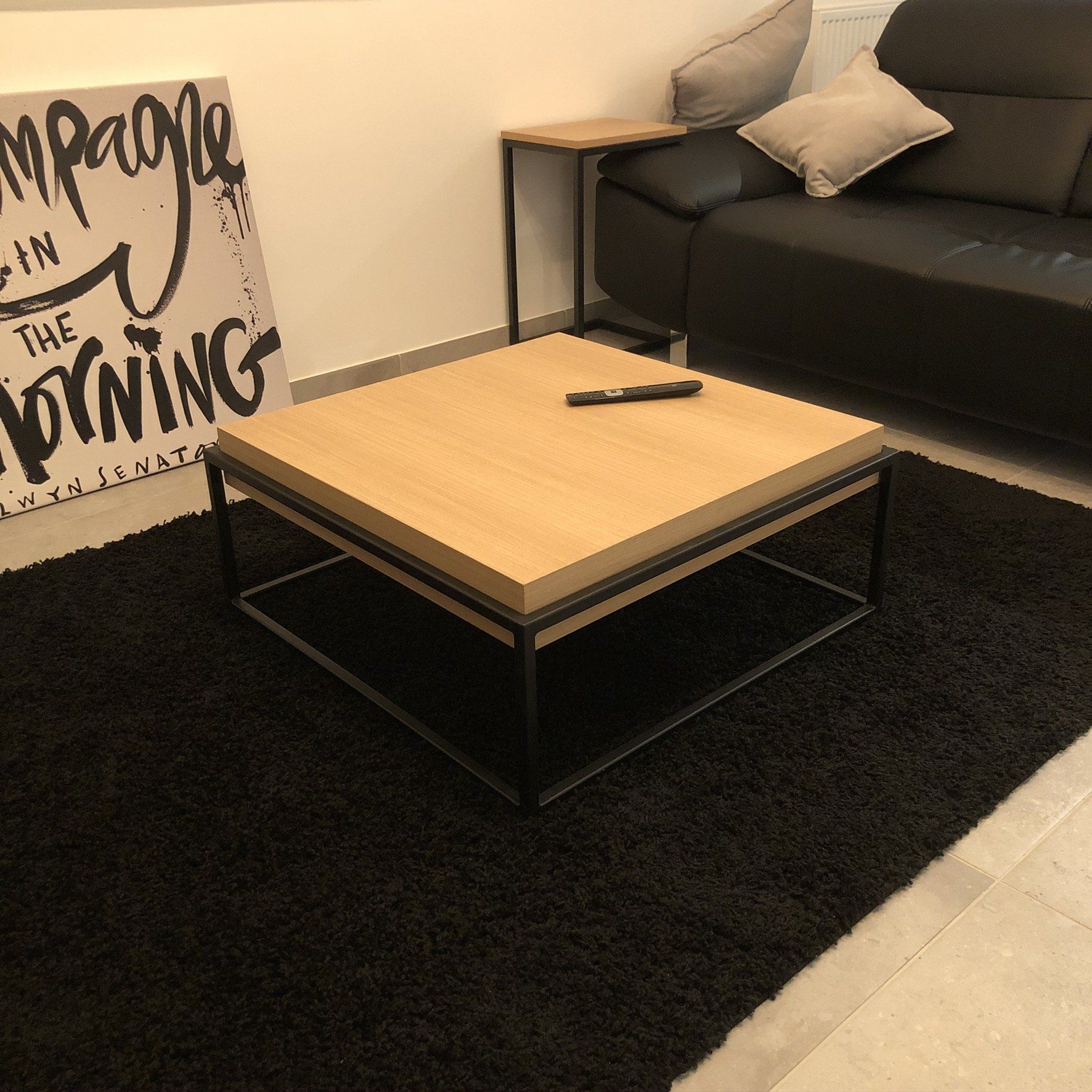 Een vierkante houten salontafel met een afstandsbediening erop, op een zwart vloerkleed, met op de achtergrond een zwarte bank.