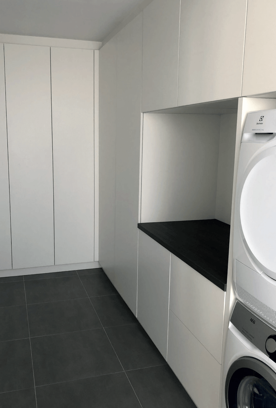 Wasruimte met witte kasten, donkere bank, wasmachine en droger. Grijze vloer.