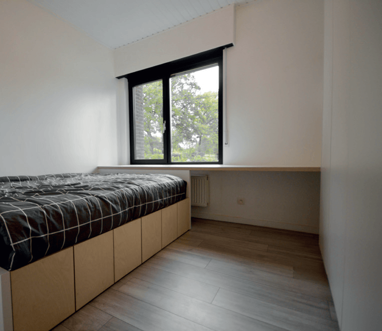 Slaapkamer met bedframe en opbergruimte. Raam met zwarte jaloezieën, houten bureau.