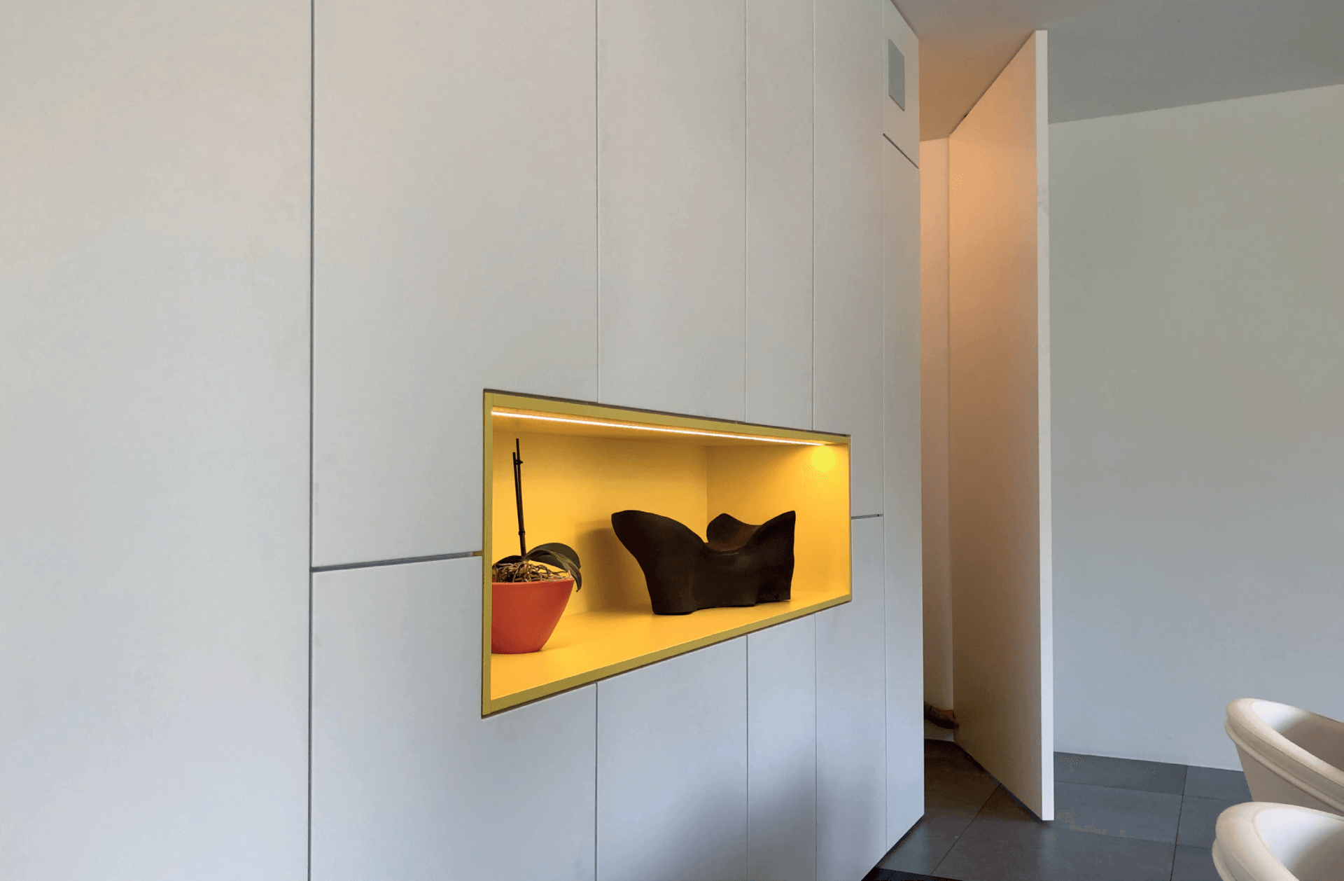 Ingebouwde muurnis met gele verlichting. Er staat een potplant en een donker beeldhouwwerk in.