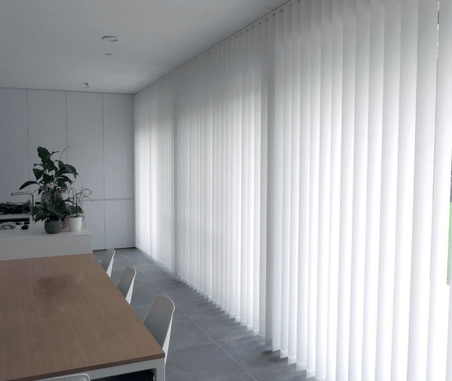 In een moderne kamer met eettafel en planten bedekken witte verticale jaloezieën een wand met ramen.