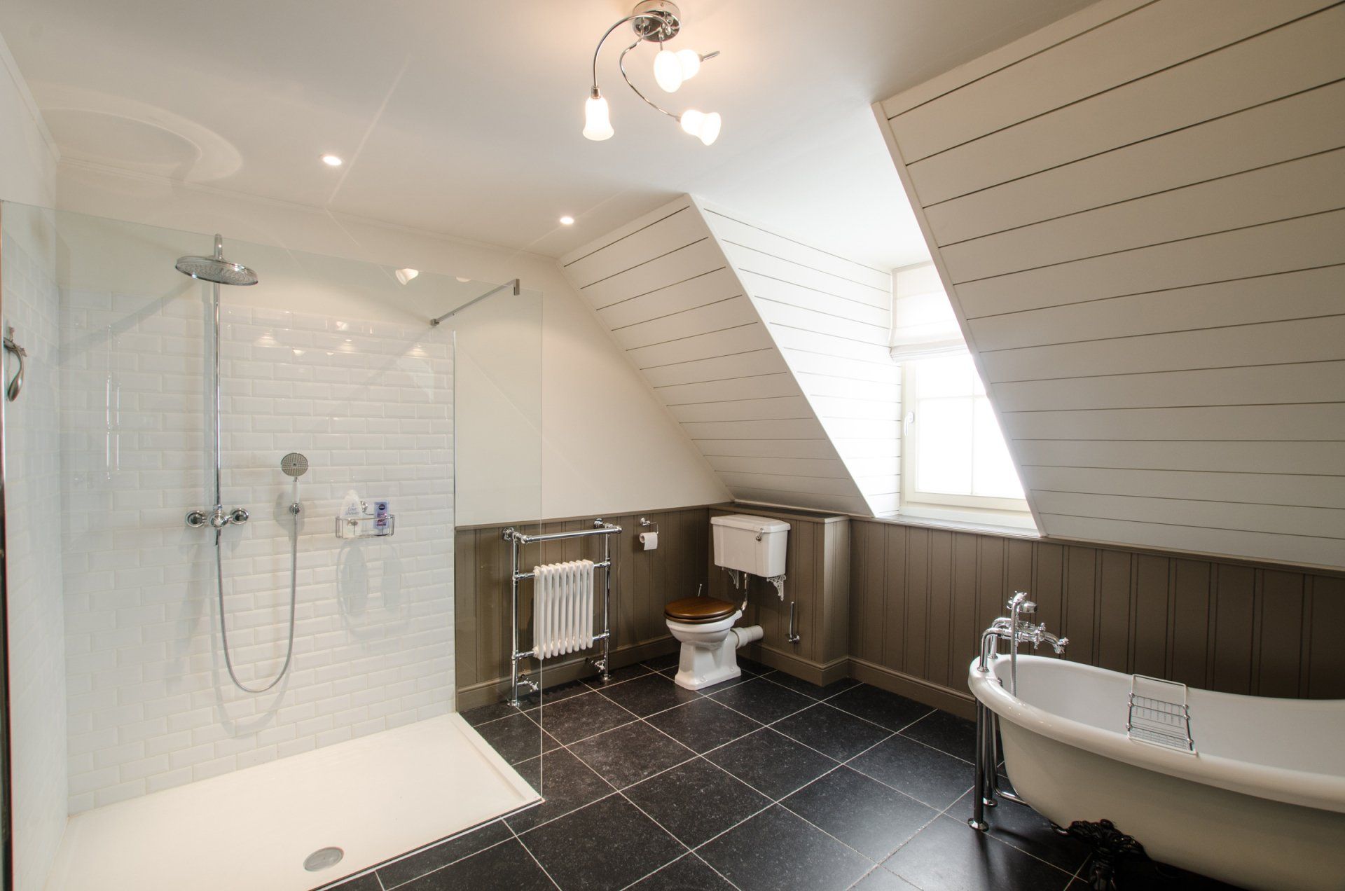 Ruime badkamer met glazen douchecabine, vrijstaand bad op pootjes en toilet onder een schuin plafond; zwart-witte tegels.