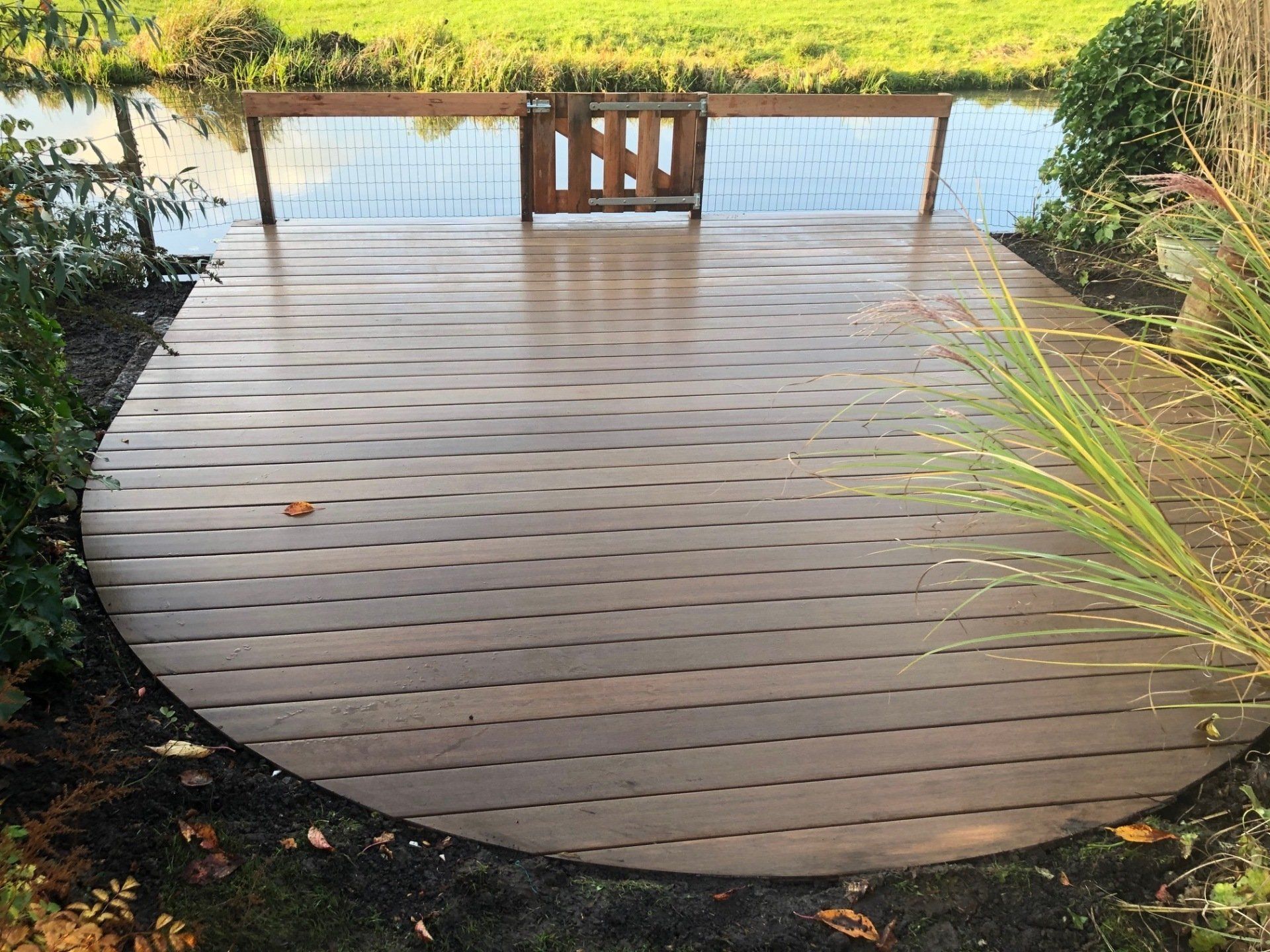 Houten terras met gebogen rand aan het water, klein poortje op de achtergrond.