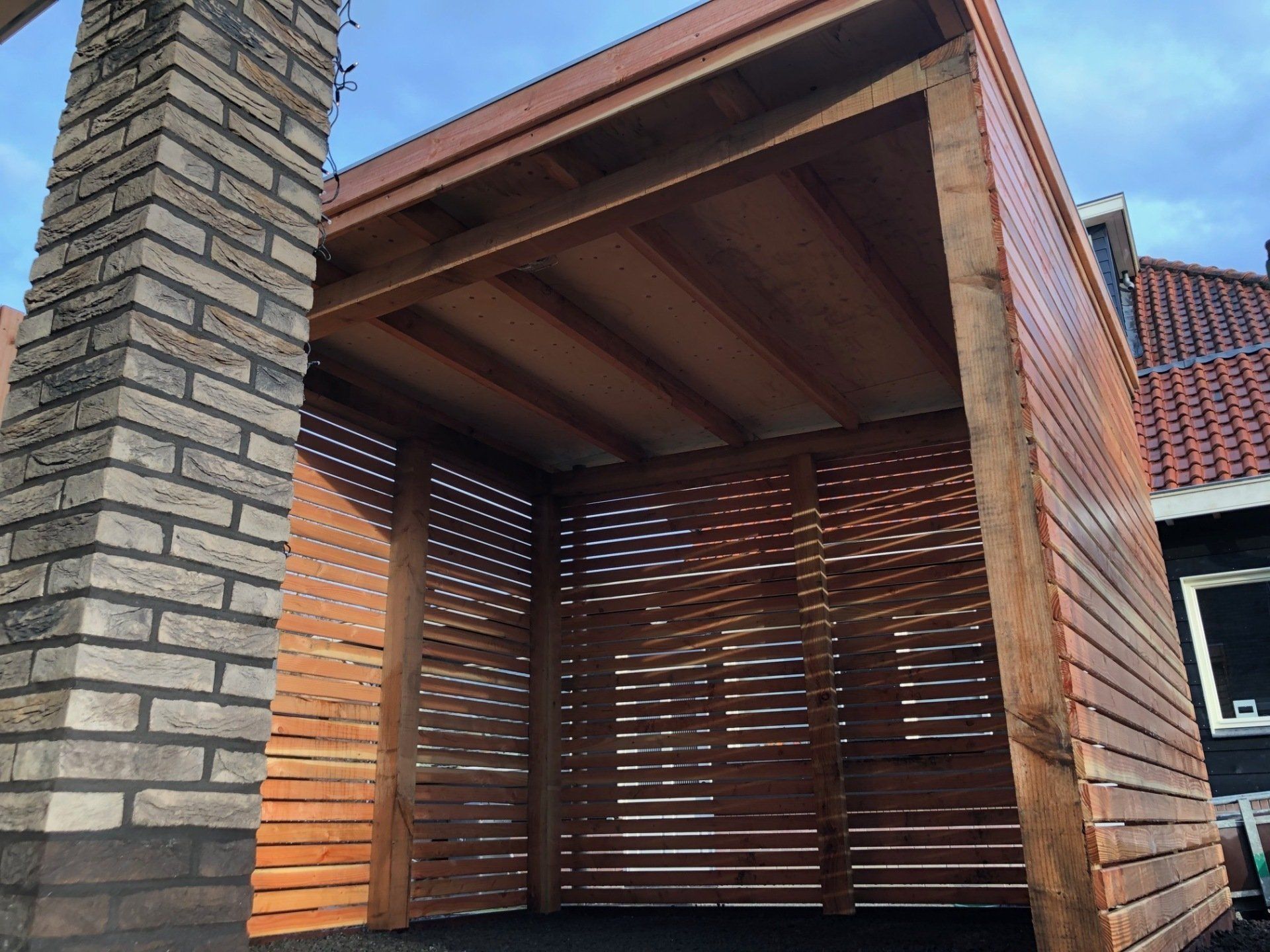 Houten tuinhuisje met lattenwanden en een bakstenen pilaar.