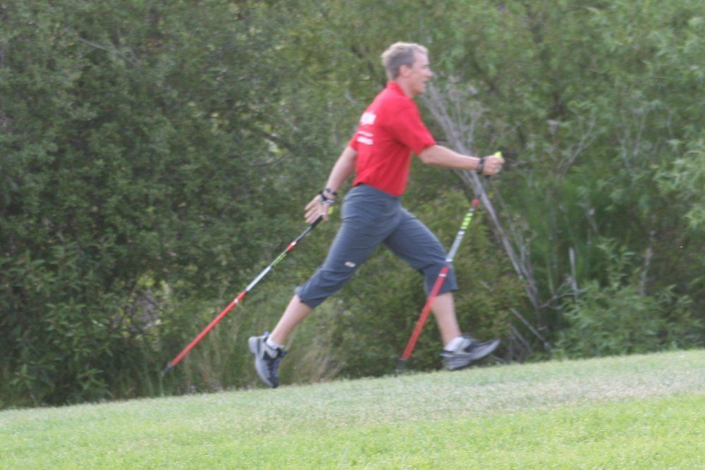 A man in a red shirt is walking on a grassy hill with walking poles.