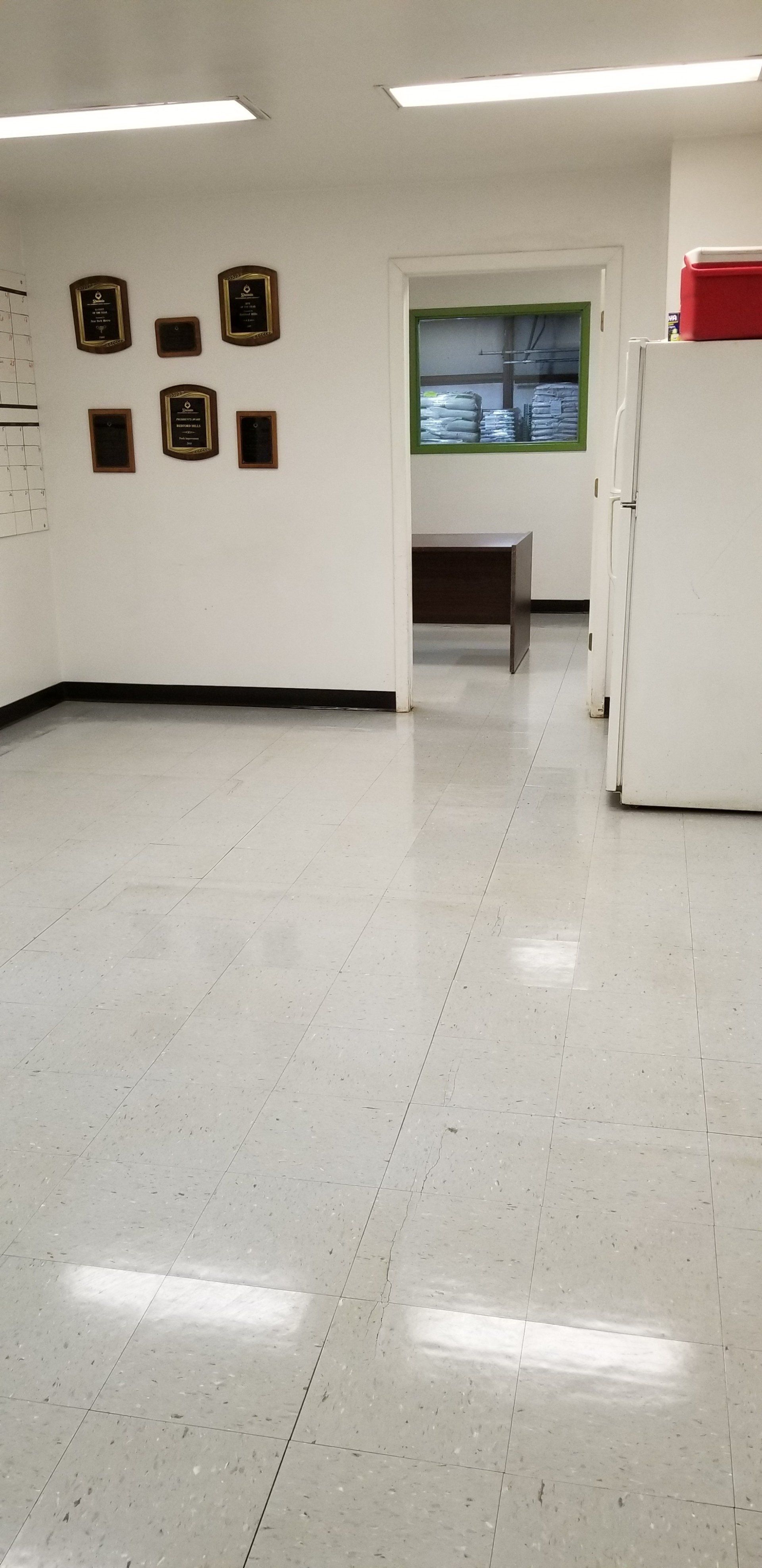 A white room with a tiled floor, several wall frames, a doorway to another office, and a white refrigerator on the right.