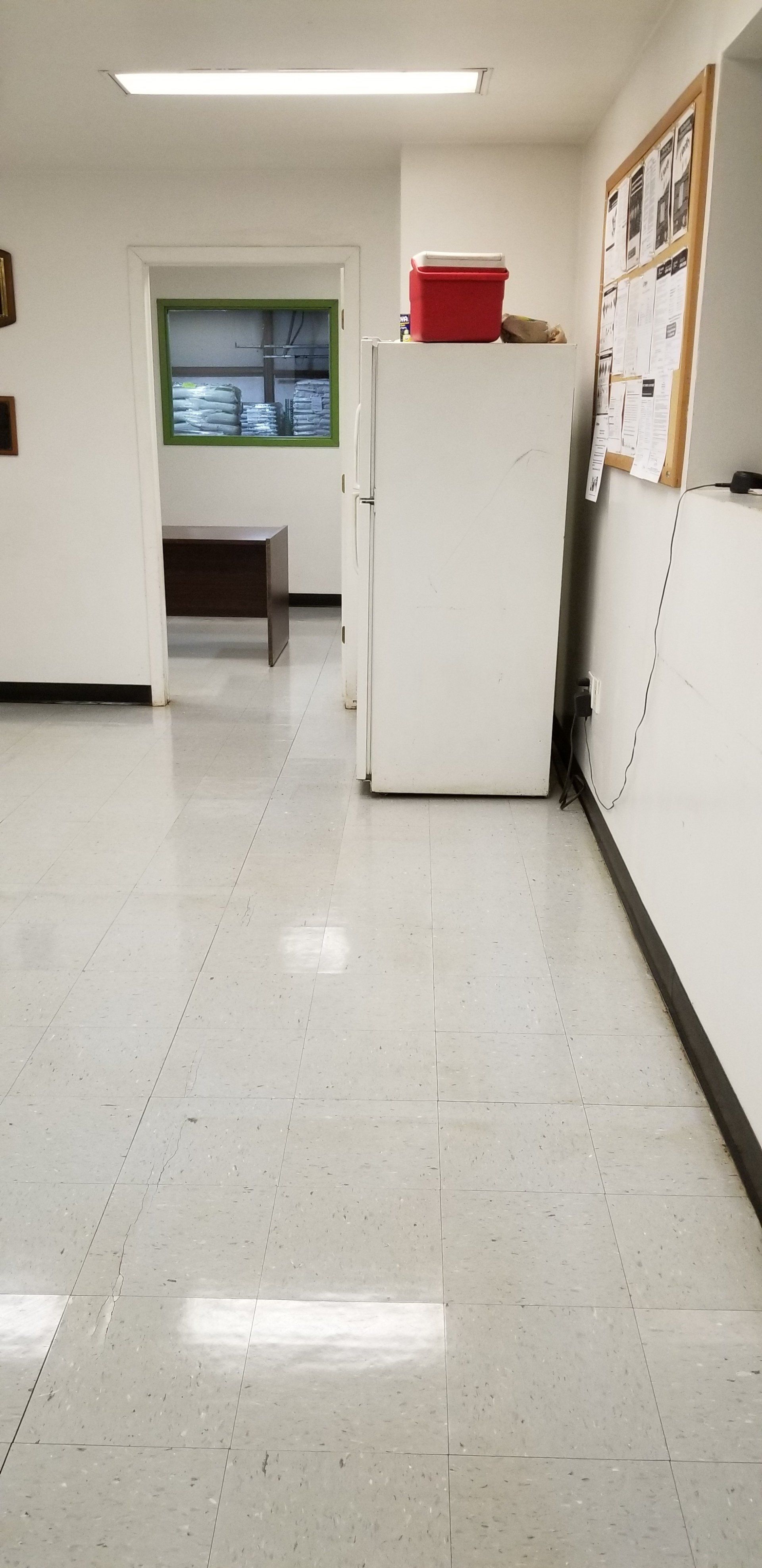 A white hallway with a tall white refrigerator, a red cooler on top, a bulletin board, and a doorway to a small office.