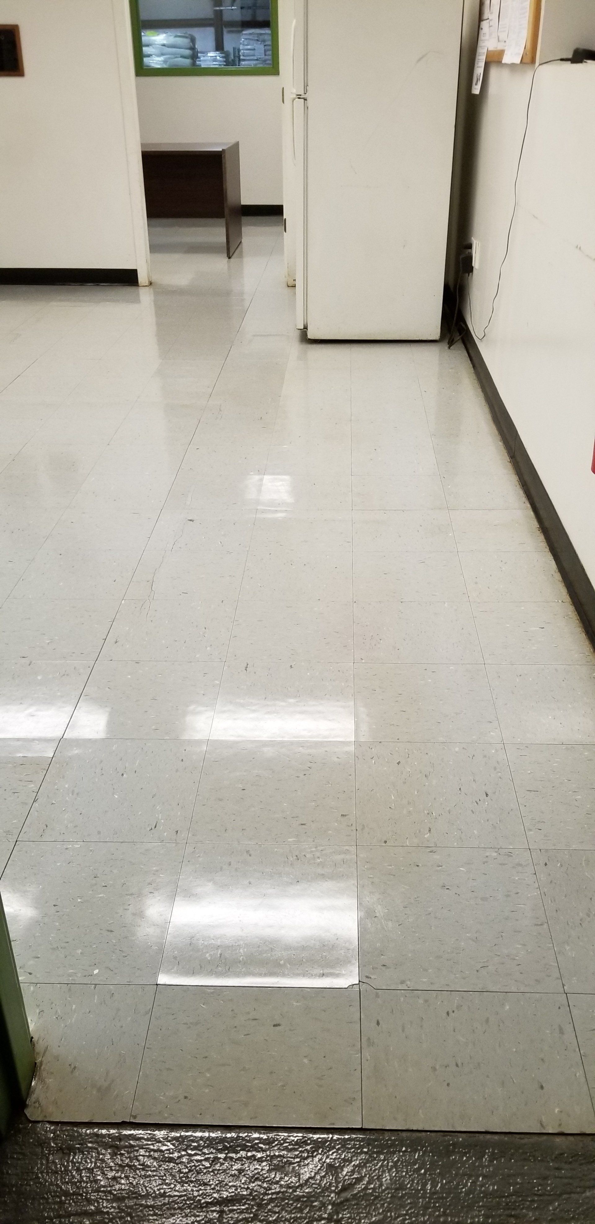 A hallway with light gray speckled vinyl tile flooring leading to a doorway with a dark desk visible in the distance.
