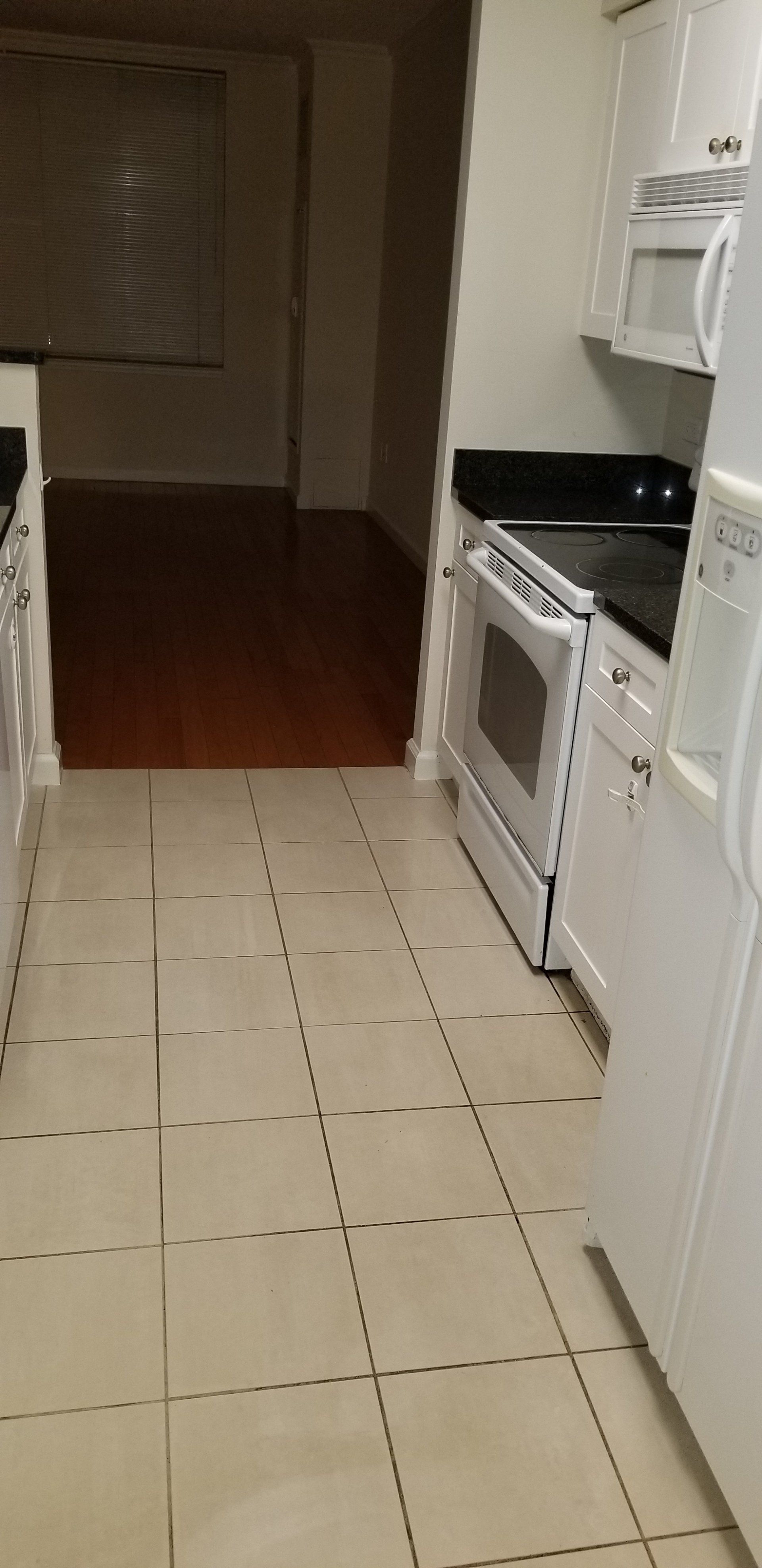 A galley-style kitchen with white cabinets, a black countertop, white appliances, and a tiled floor leading to a hallway.
