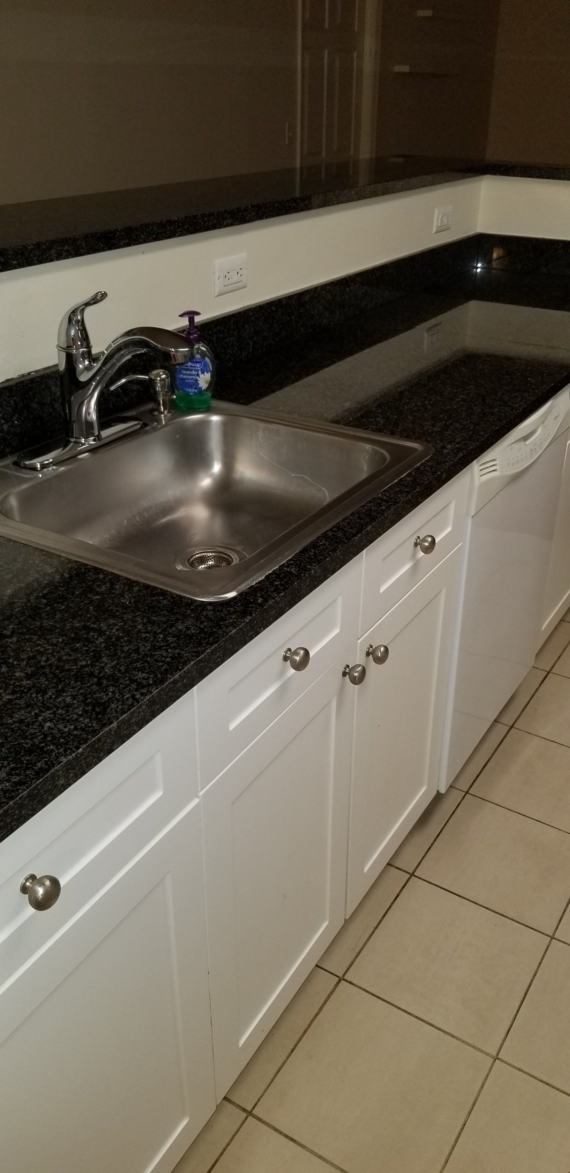 A stainless steel kitchen sink with a faucet and soap dispenser, set in a dark granite countertop above white cabinets.