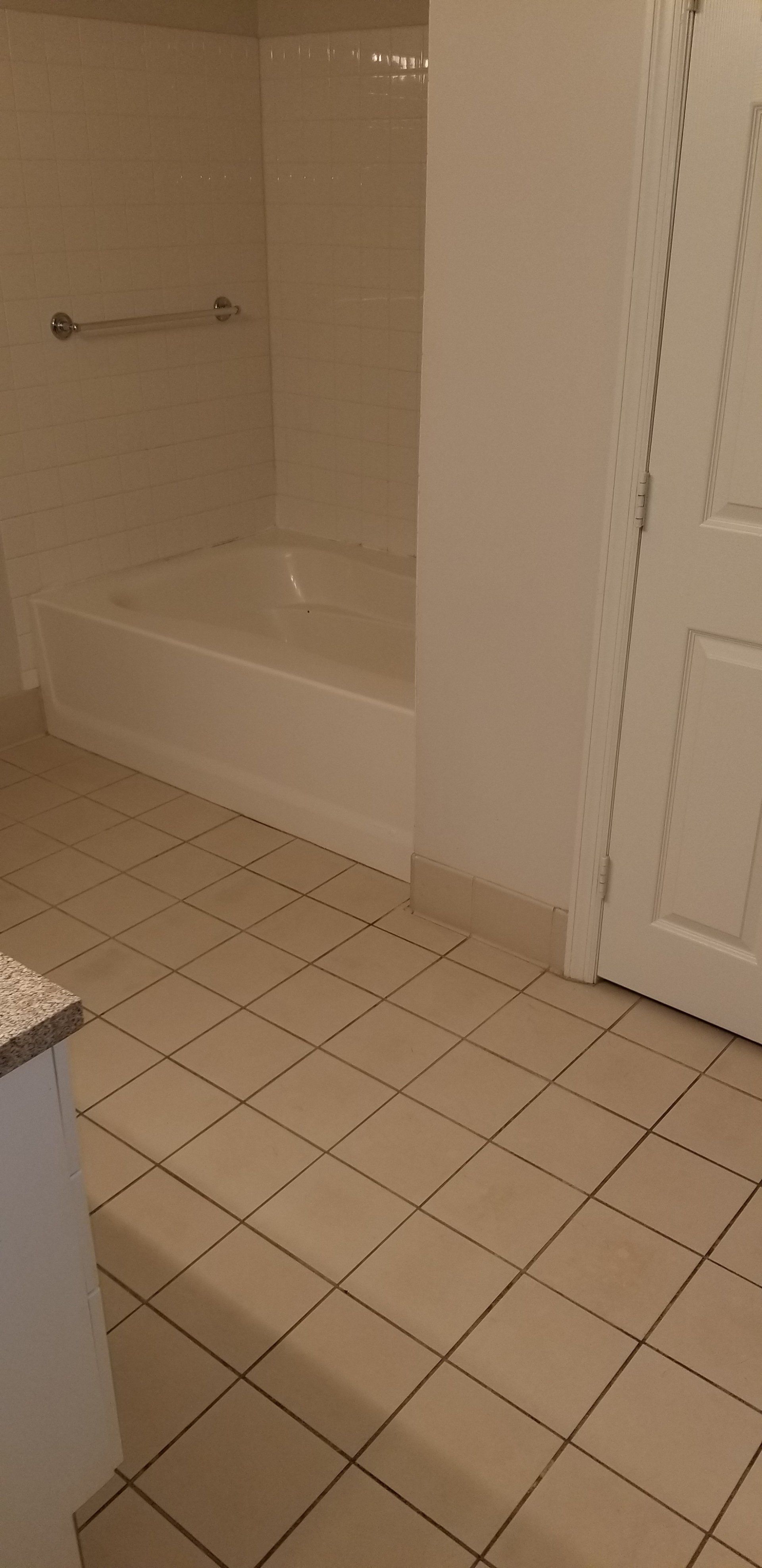 A beige-tiled bathroom featuring a white bathtub with a grab bar on the wall and a white door to the right.