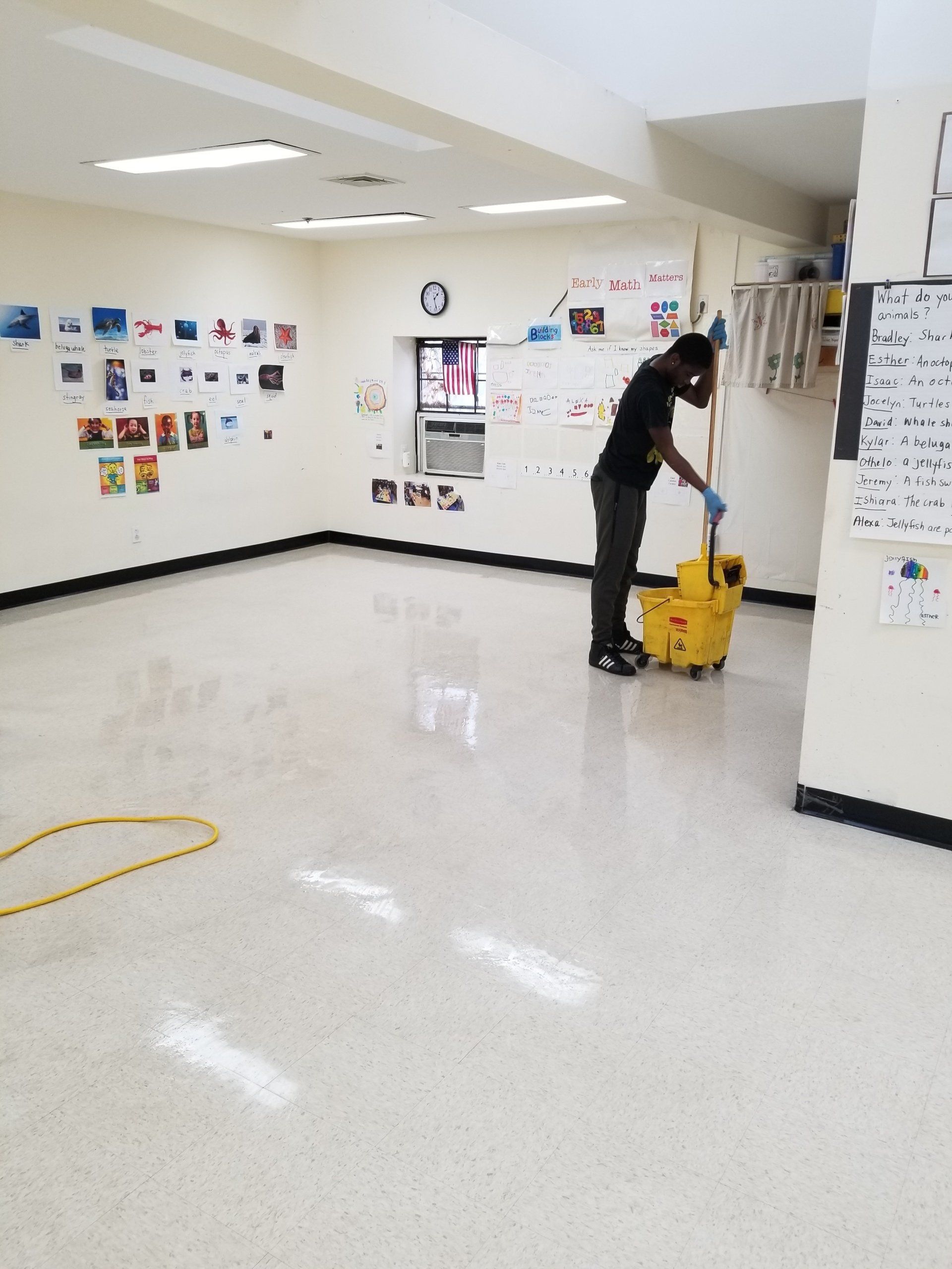 A person in a classroom uses a yellow mop bucket to clean the shiny, light-colored floor.