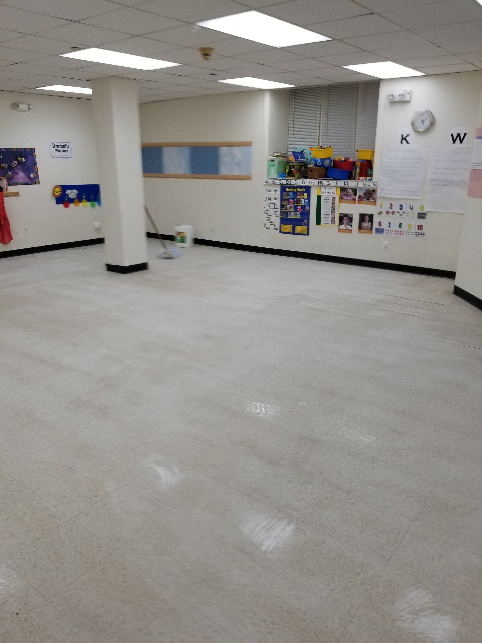 An empty, brightly lit classroom with light-colored flooring, a support pillar, and educational posters on the walls.