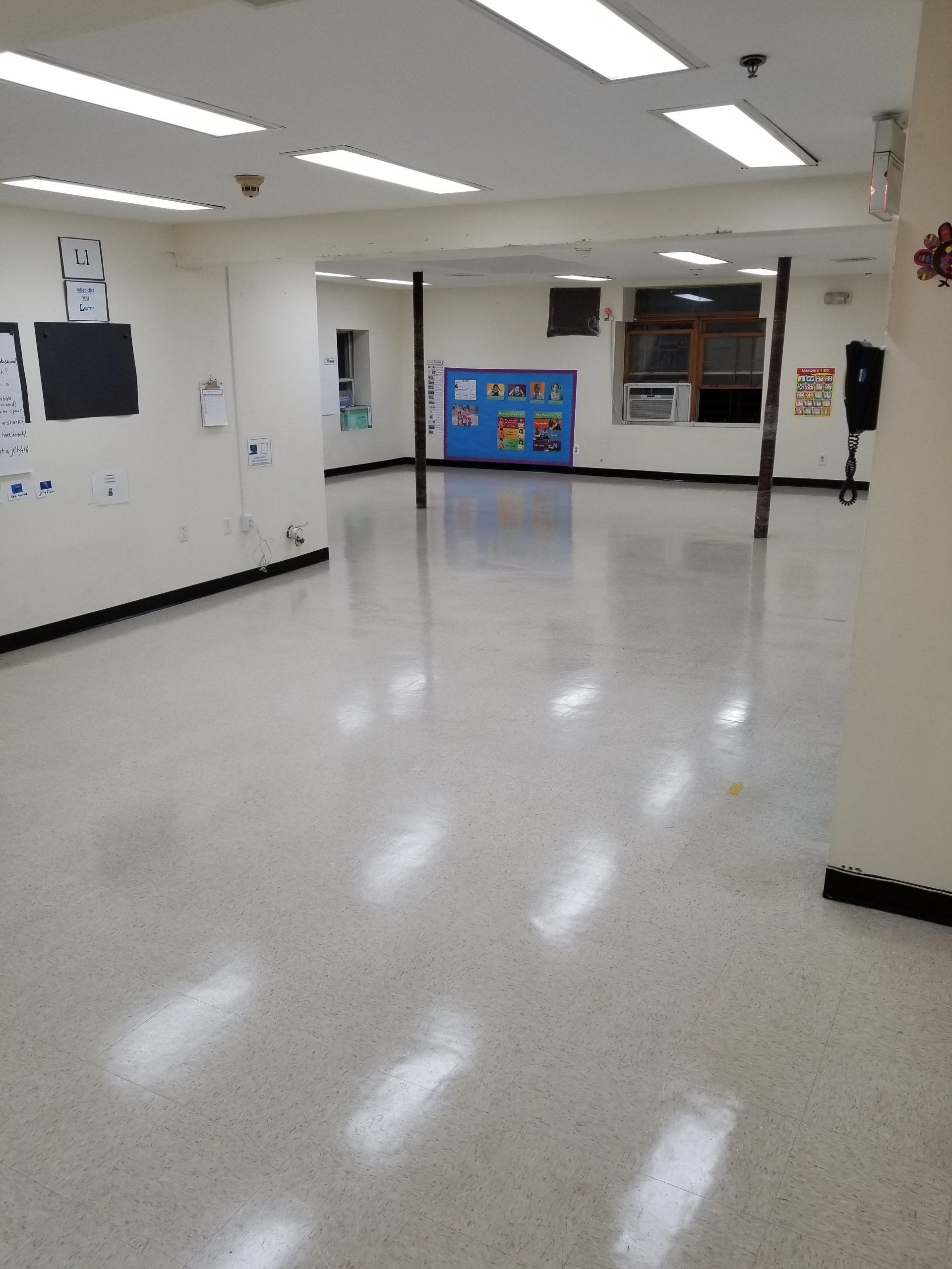 A wide, empty room with polished light-colored floors, white walls, and bright overhead fluorescent lighting.