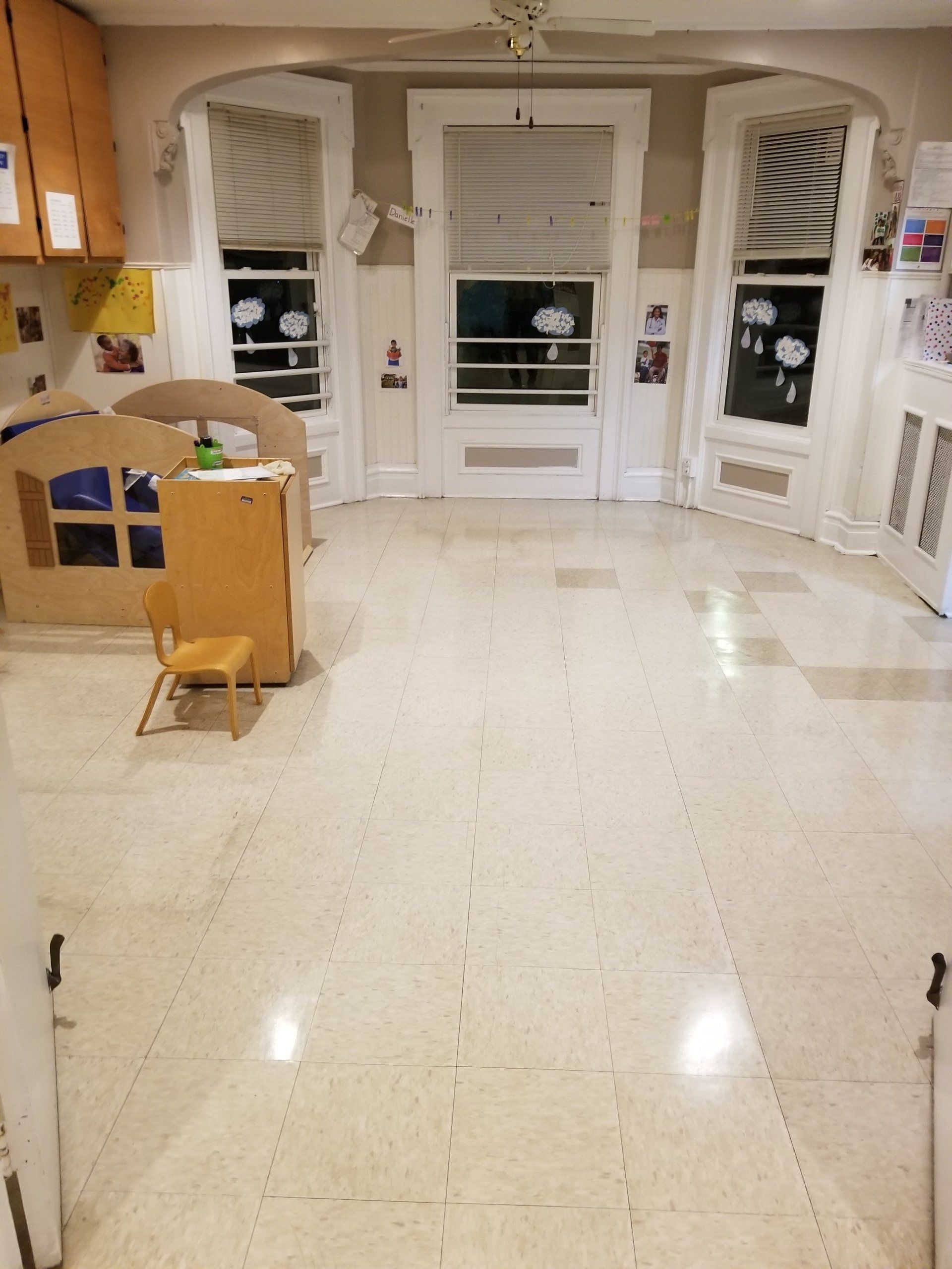 An indoor room with beige tile flooring, three large windows, a small wooden chair, and child-sized play equipment.