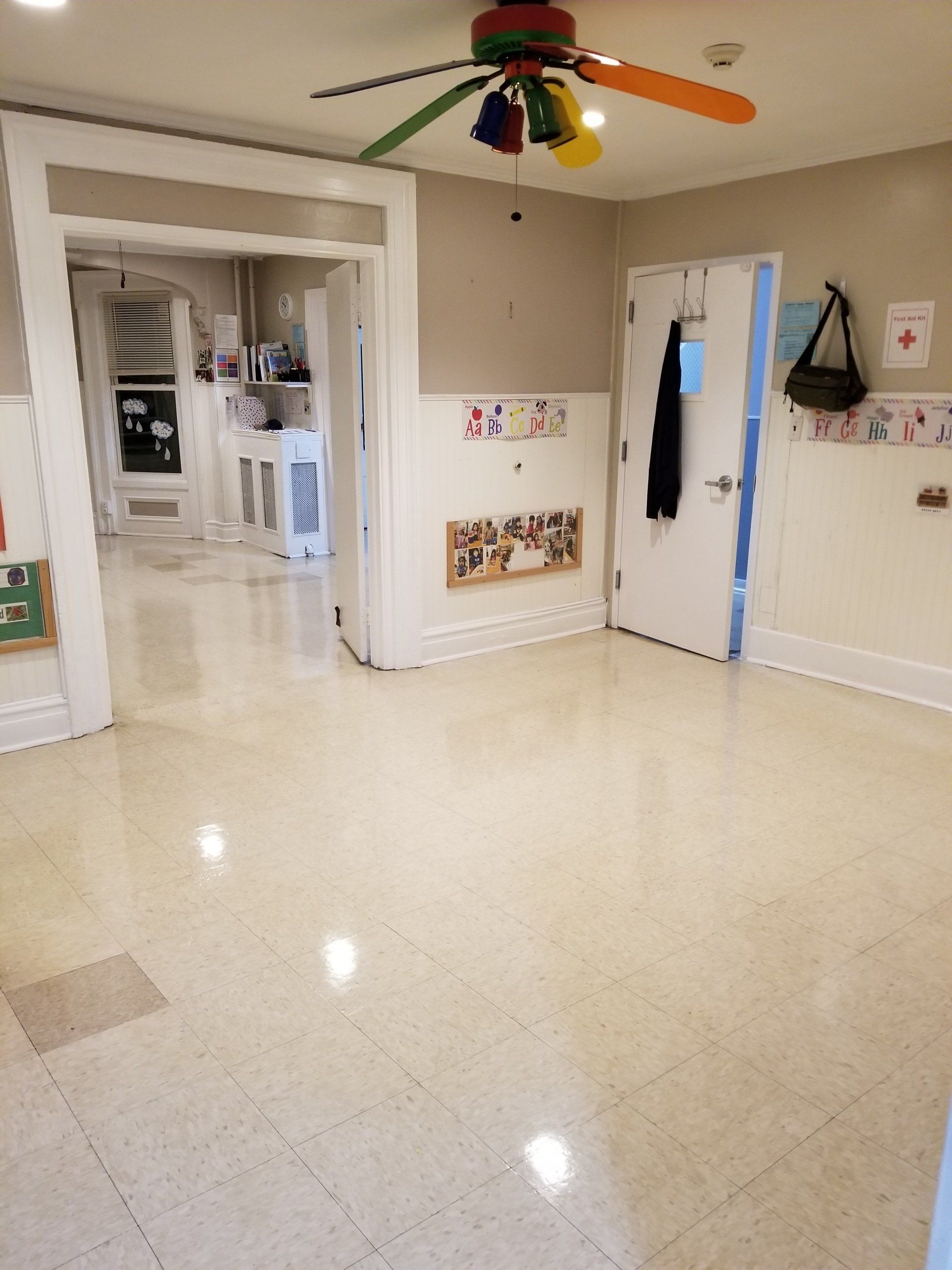 Brightly lit school room hallway with beige tile floors, white walls, and a colorful, multi-blade ceiling fan.