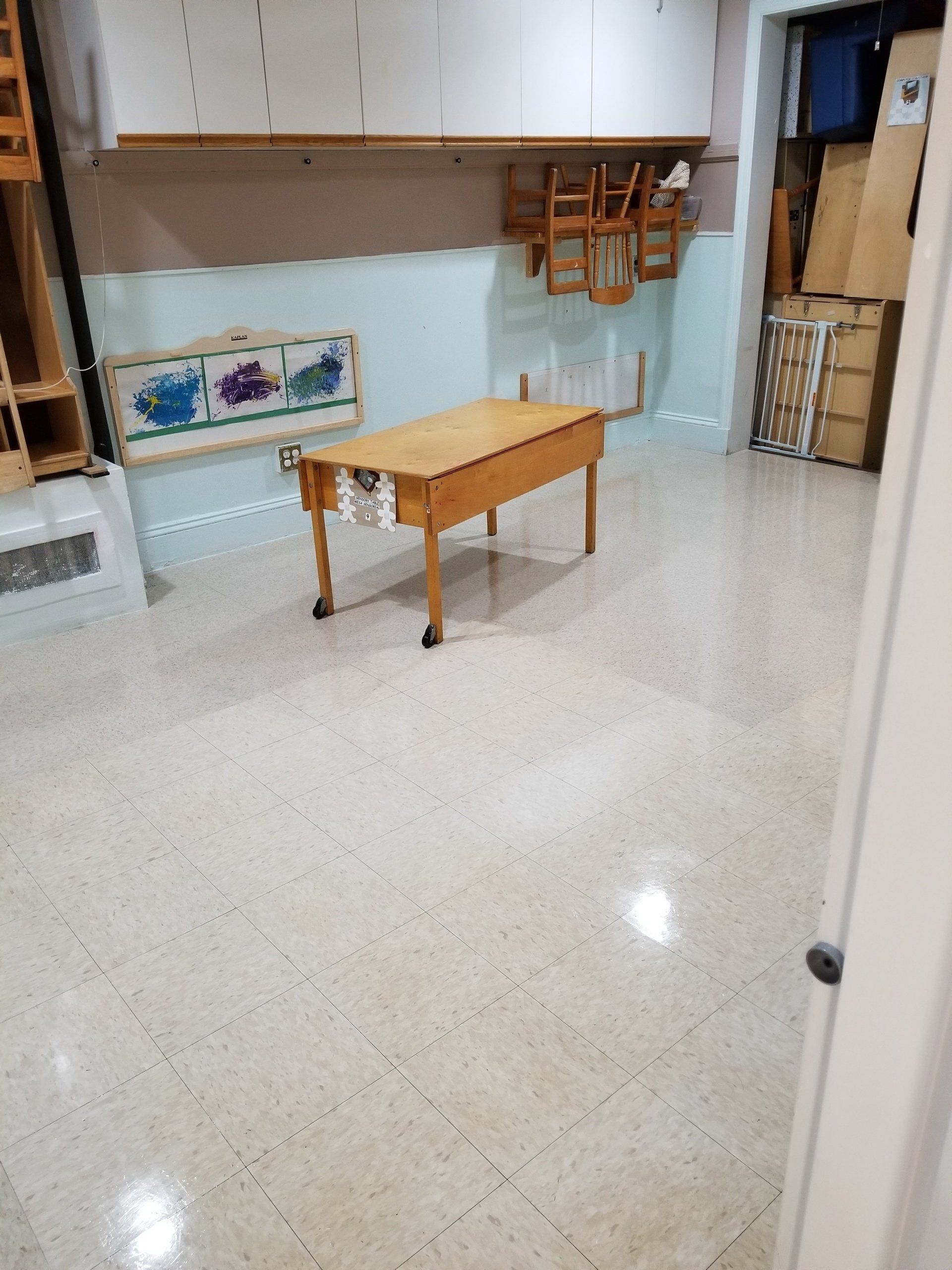 A light-colored room with a wooden drop-leaf table on wheels, wall-mounted storage, and chairs hanging on the wall.
