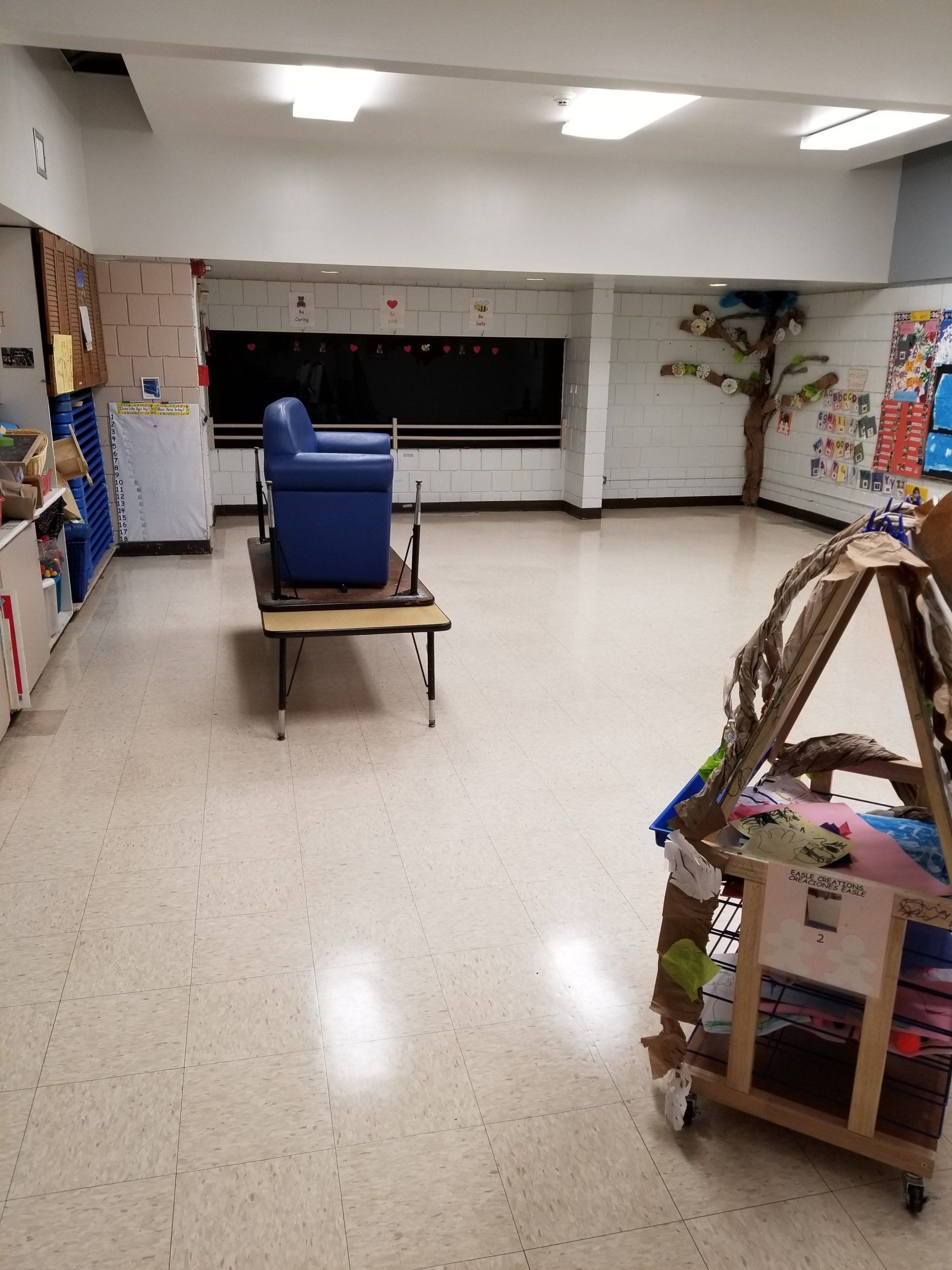 An indoor classroom with a blue chair placed on a small table and a wooden storage cart in the foreground.
