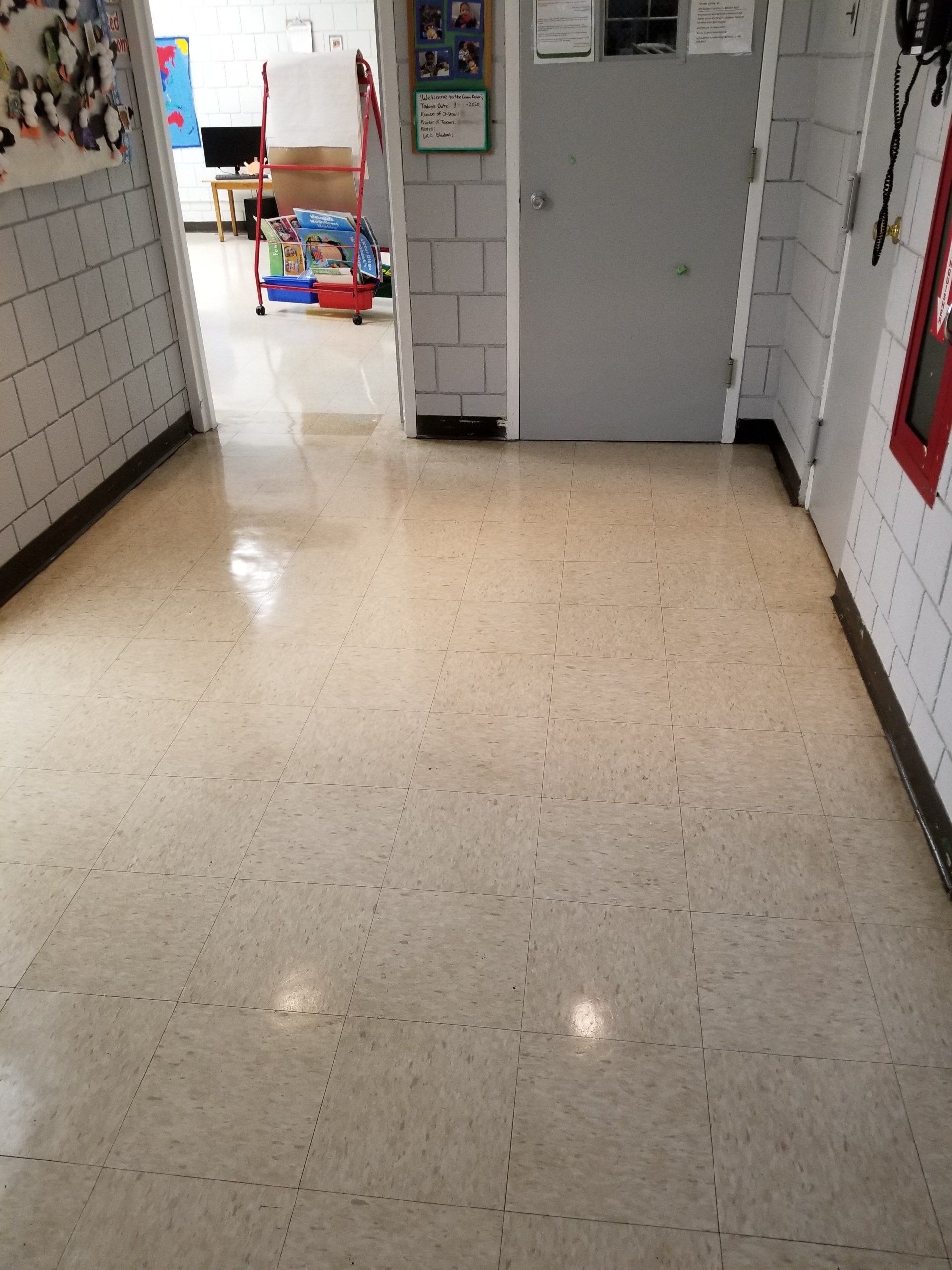 A view down a school hallway with light-colored tile floors, a gray door, and a partially open doorway in the background.