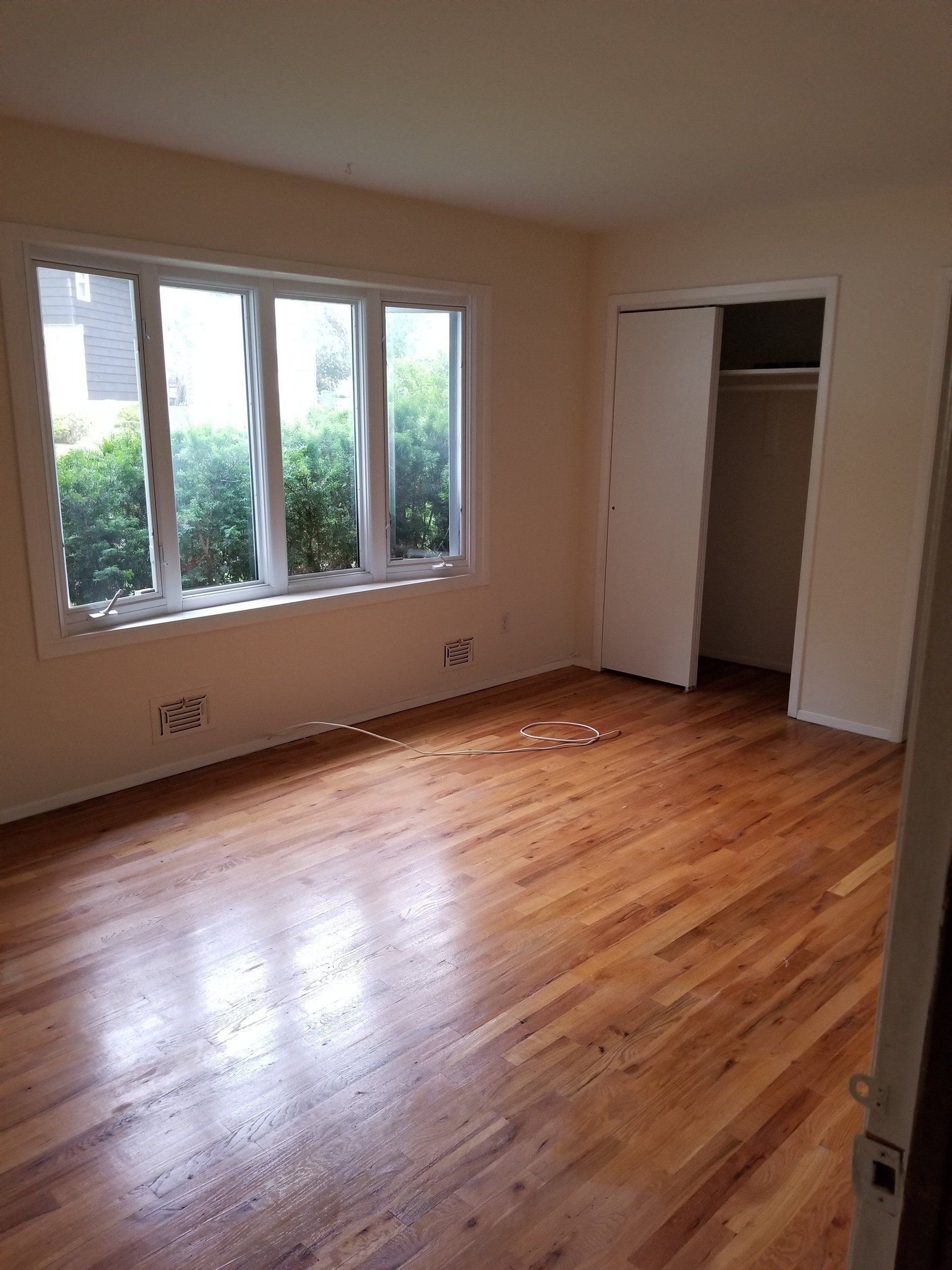 An empty room with polished hardwood floors, cream walls, a large window looking onto greenery, and an open closet.