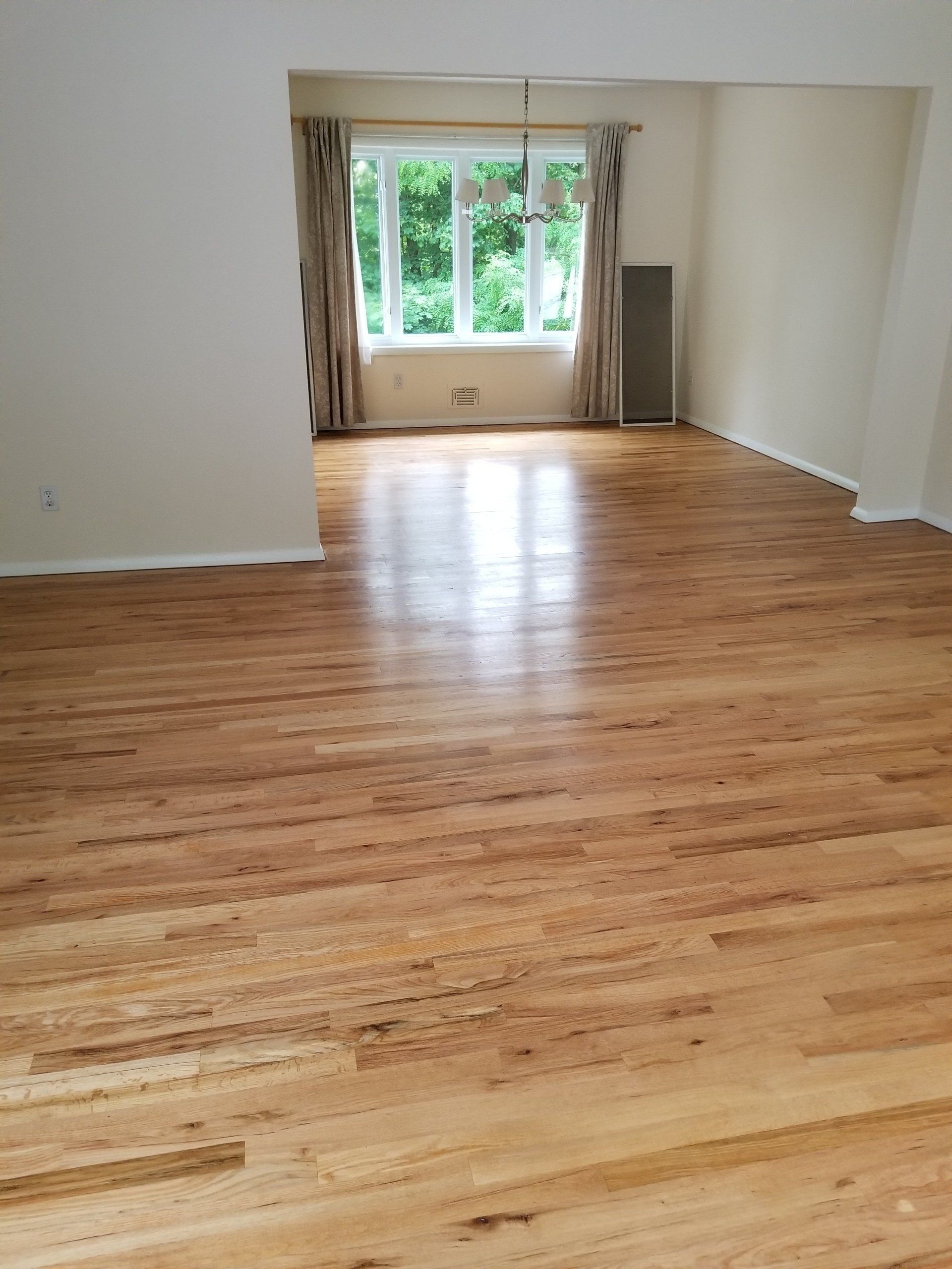 A spacious room with polished light wood flooring, beige walls, and a large window with drapes in the background.