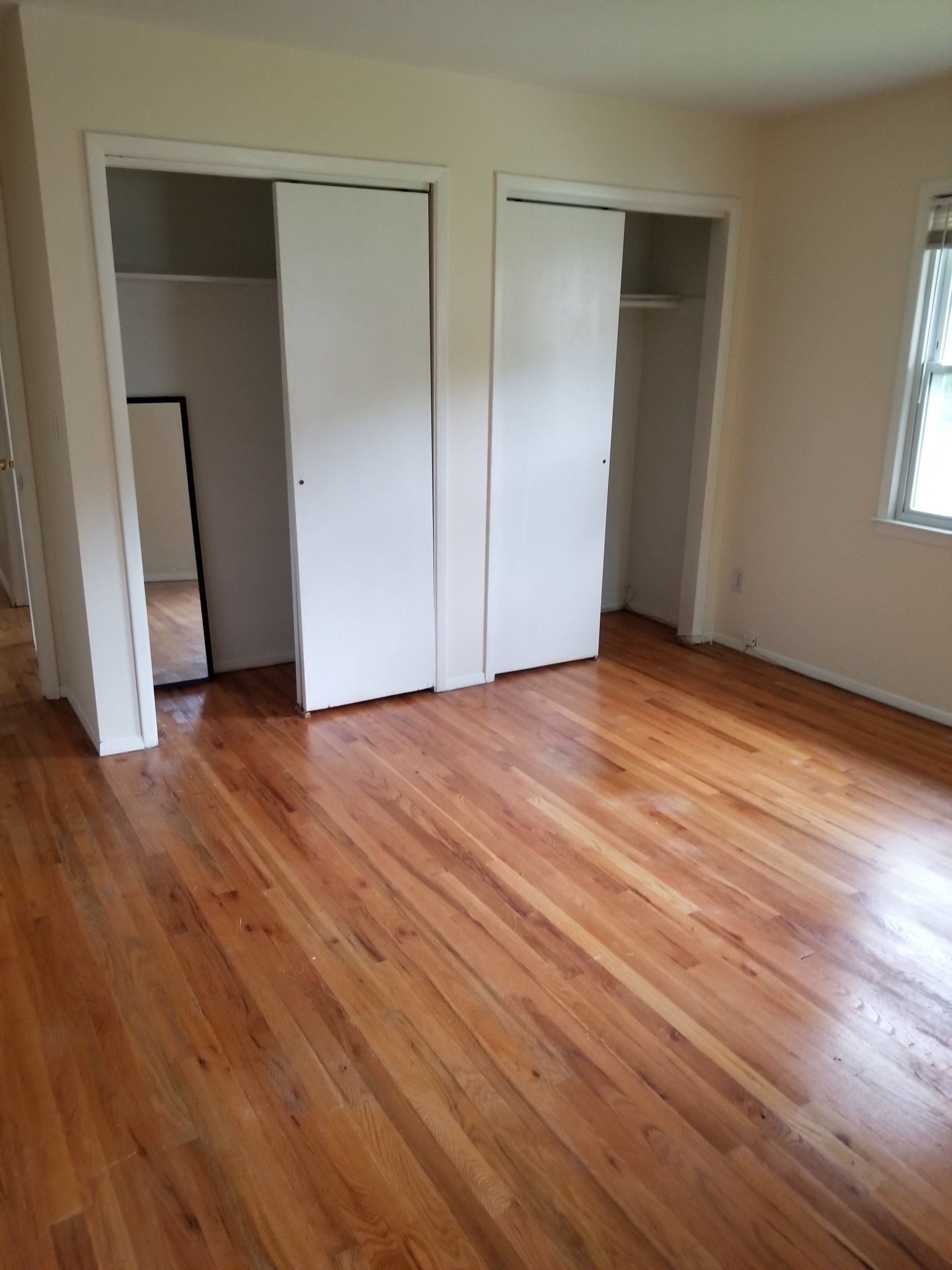 An empty room with polished hardwood floors, cream walls, two white closet doors, and a window on the right.
