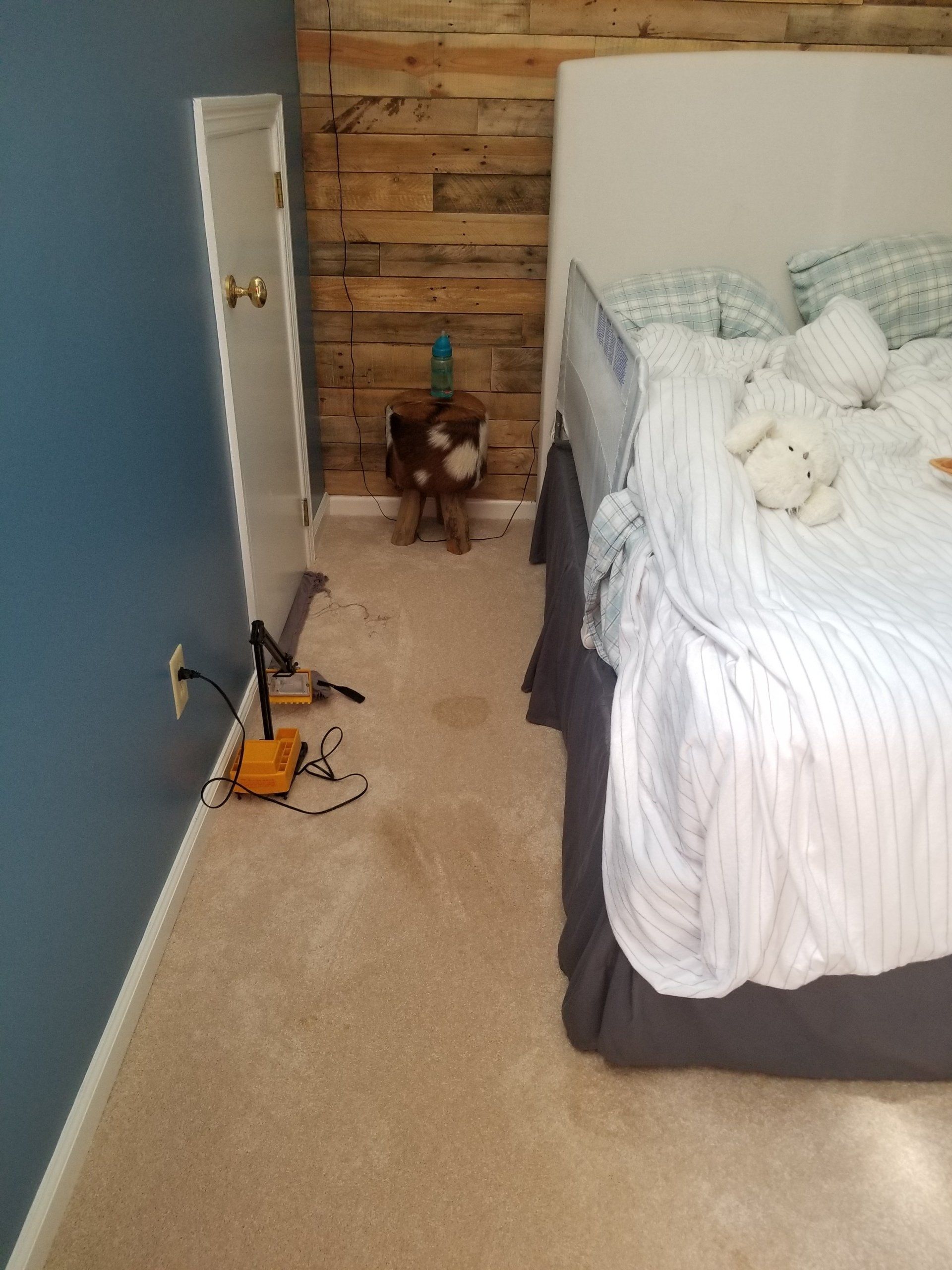 A bedroom corner with a blue wall, wood-paneled wall, a bed with a white comforter, and a small, cow-print stool.