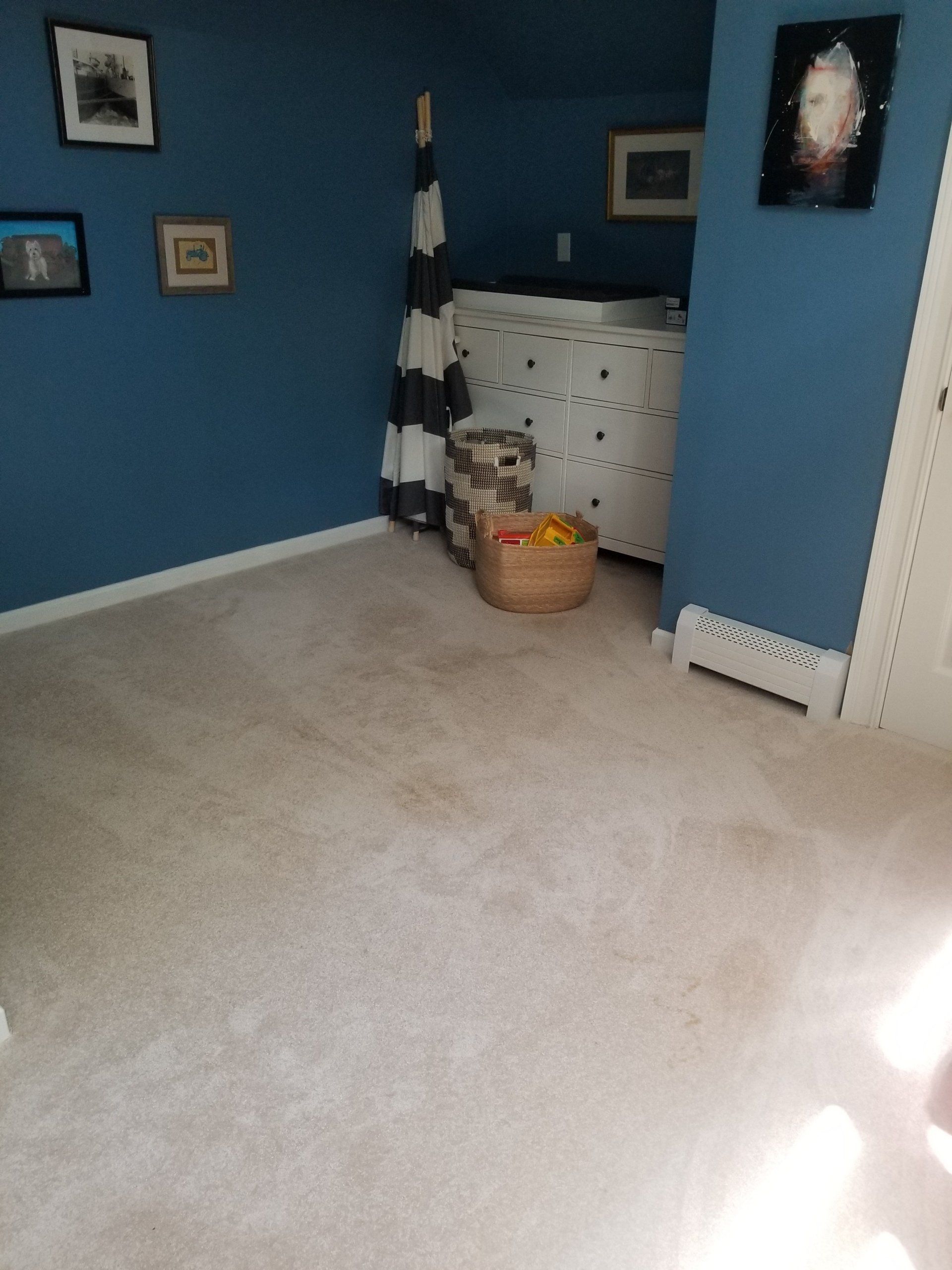 A blue-walled room with beige carpet, a white dresser, a patterned floor cushion, a storage bin, and wall-mounted art.