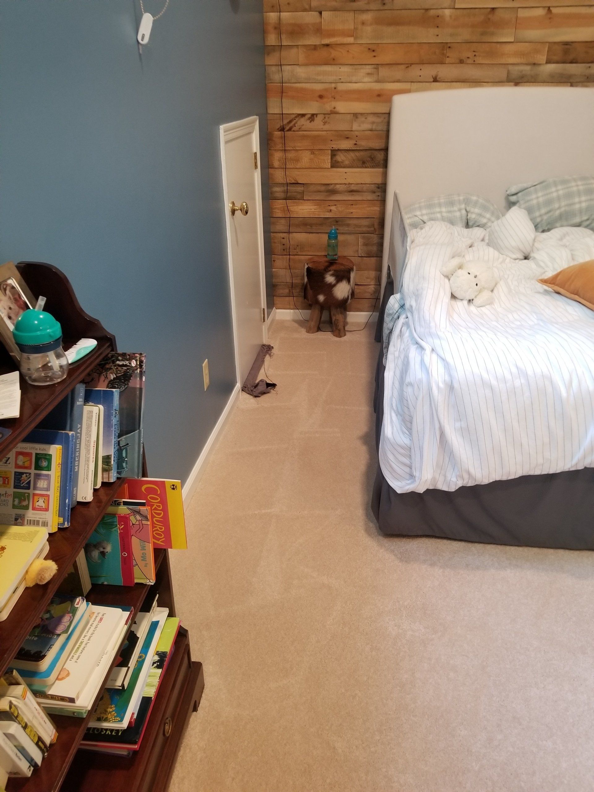 A bedroom corner with a wooden plank accent wall, a white bed, a small storage door, and a bookshelf filled with books.