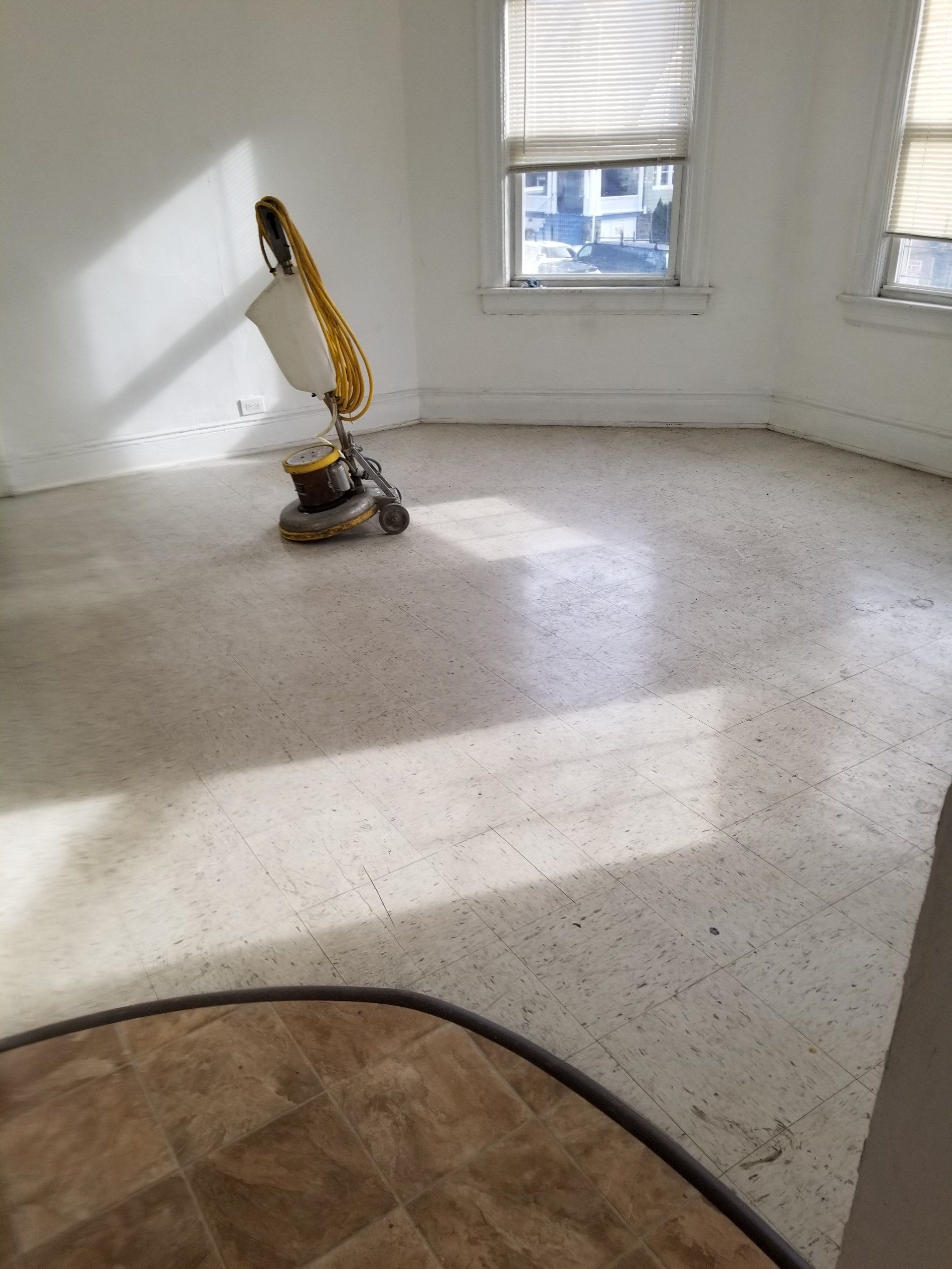 A floor buffer sits on a white, speckled tiled floor in a bright, sunlit room with windows.