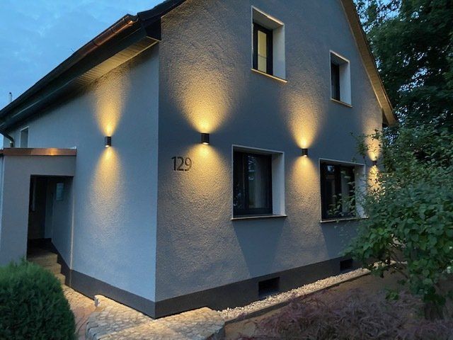 Graues Haus mit moderner Beleuchtung, die die Fassade erhellt; die Hausnummer ist 129.