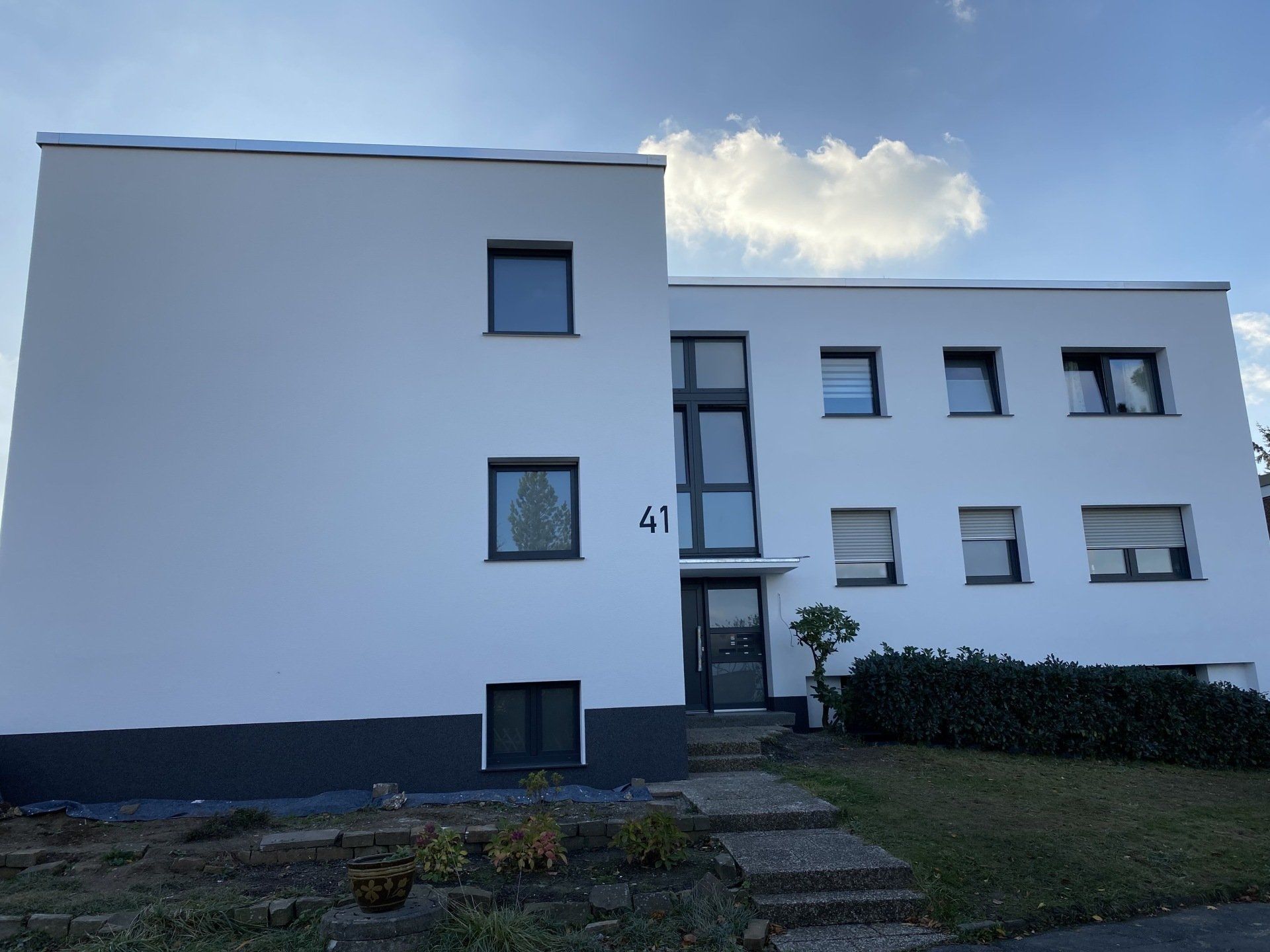Weißes zweistöckiges Gebäude mit dunkelgrauen Verzierungen, Nummer 47 über der Tür, blauer Himmel und ein kleiner Garten.