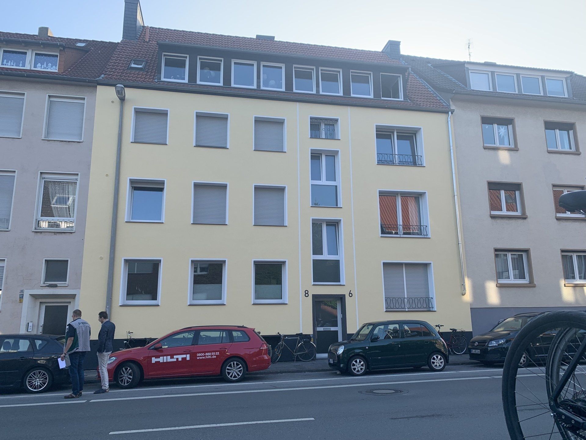 Ein gelbes Wohnhaus mit geparkten Autos und zwei Personen, die auf dem Bürgersteig stehen.