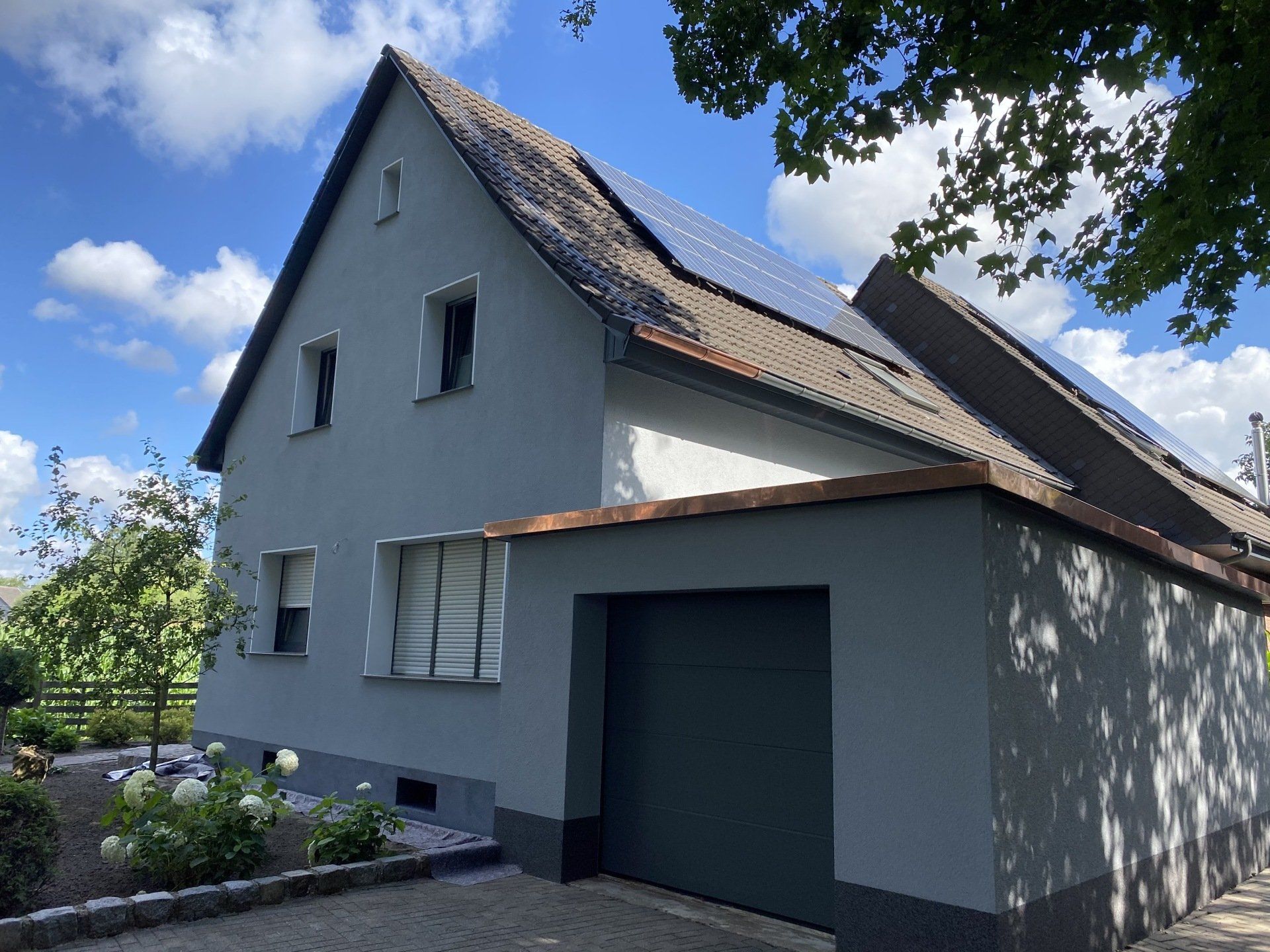Graues Haus mit Solarzellen auf dem Dach, Garage und blauem Himmel.