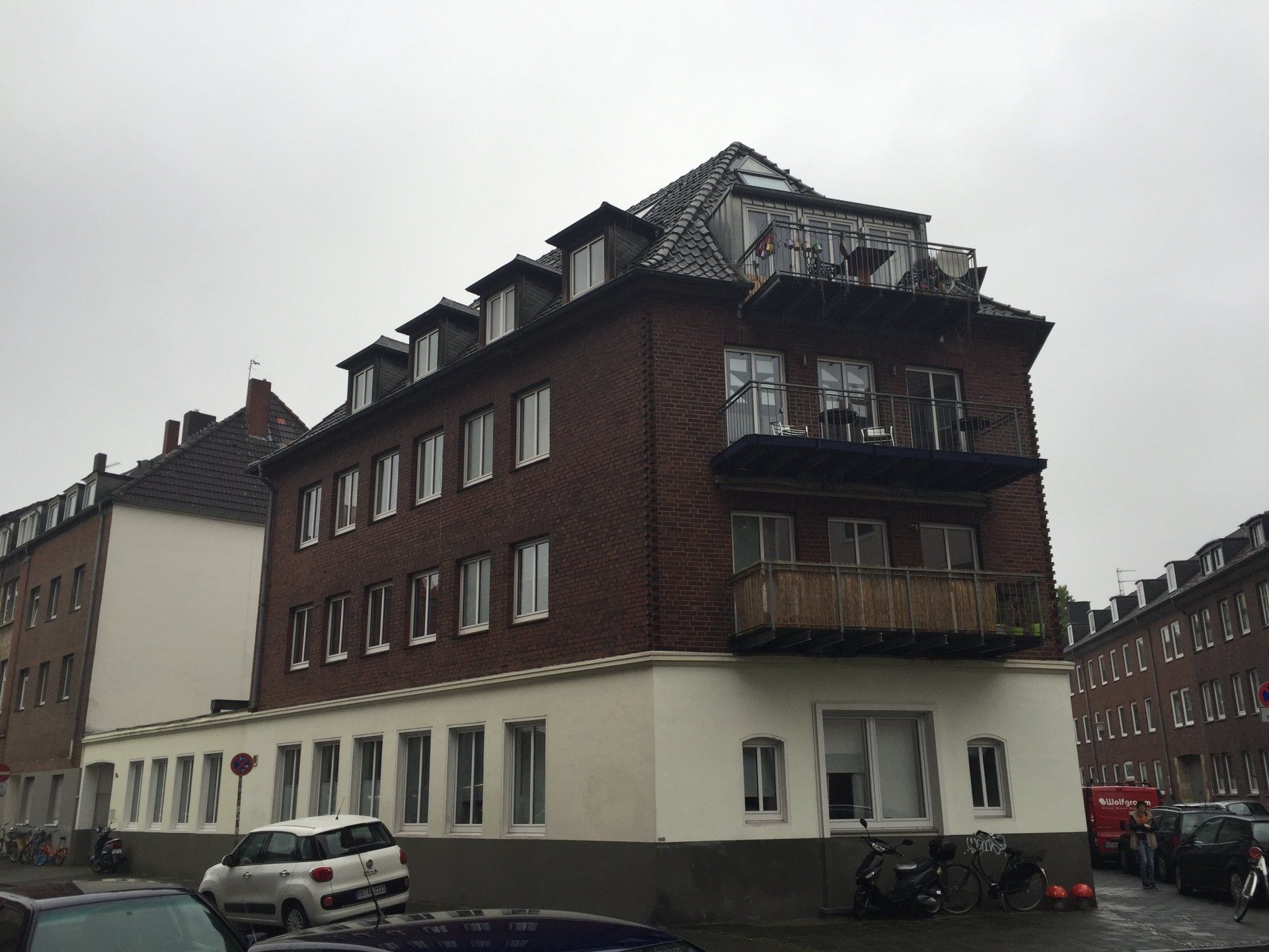 Rotes Backsteingebäude mit weißem Sockel, Balkonen und Dachgauben an einem bewölkten Tag.