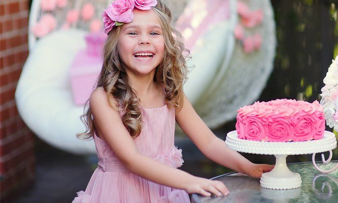 Smiling child in a pink dress beside a pink frosted cake with a floral headband