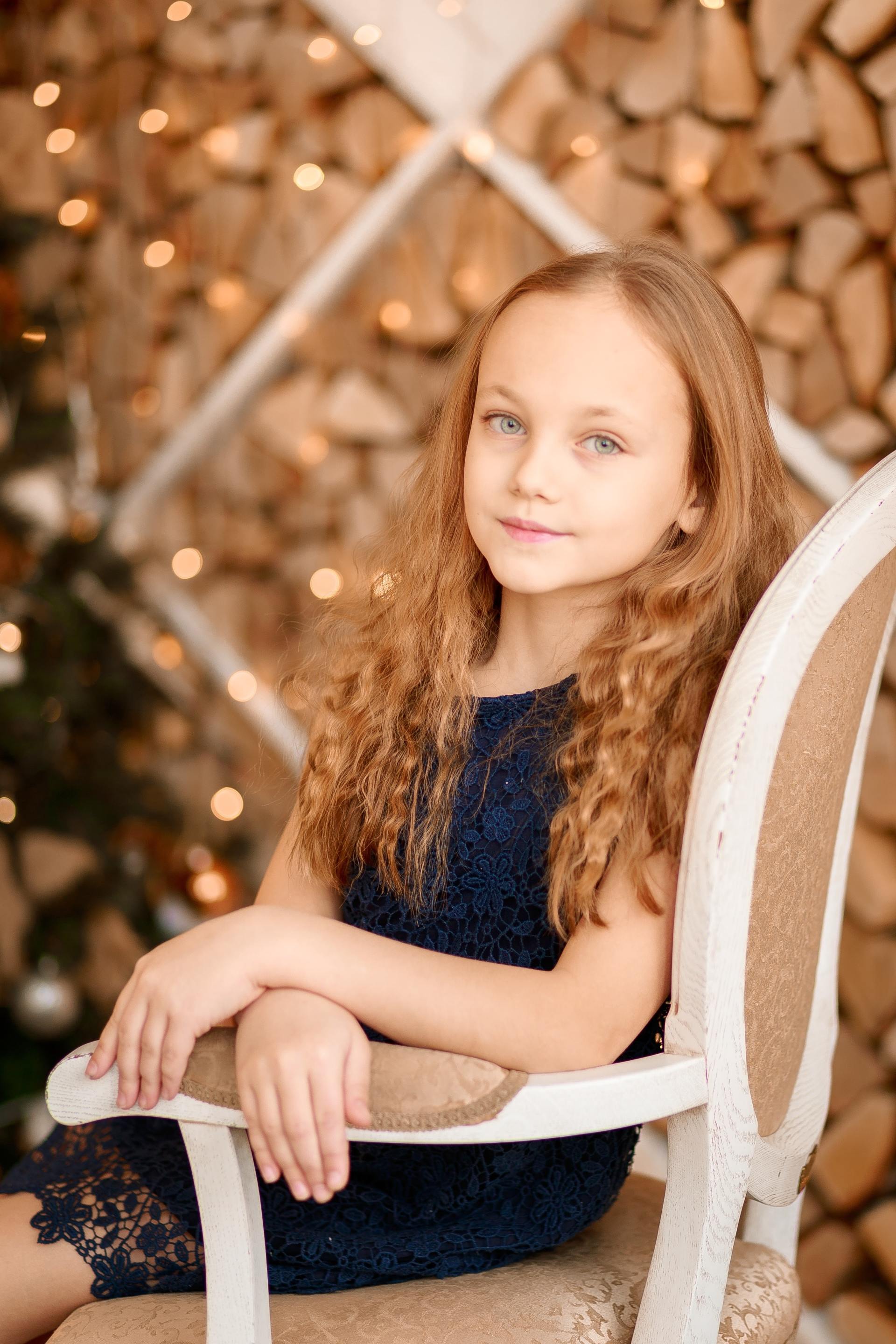 Young child sitting on a white chair, with curly hair, against a warm wood and string-light background