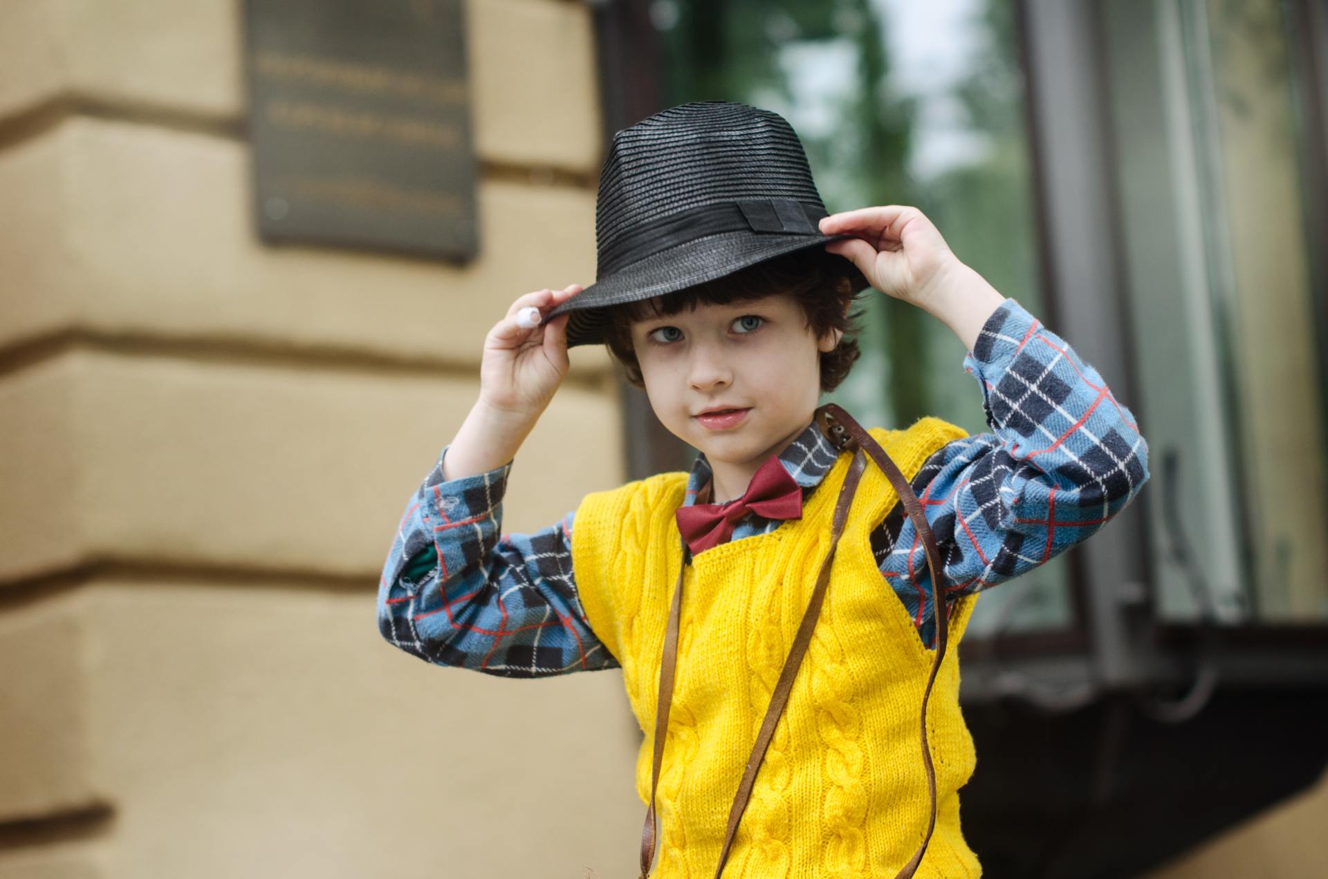 Child in a yellow vest adjusting a black hat outdoors, with a train in the background.
