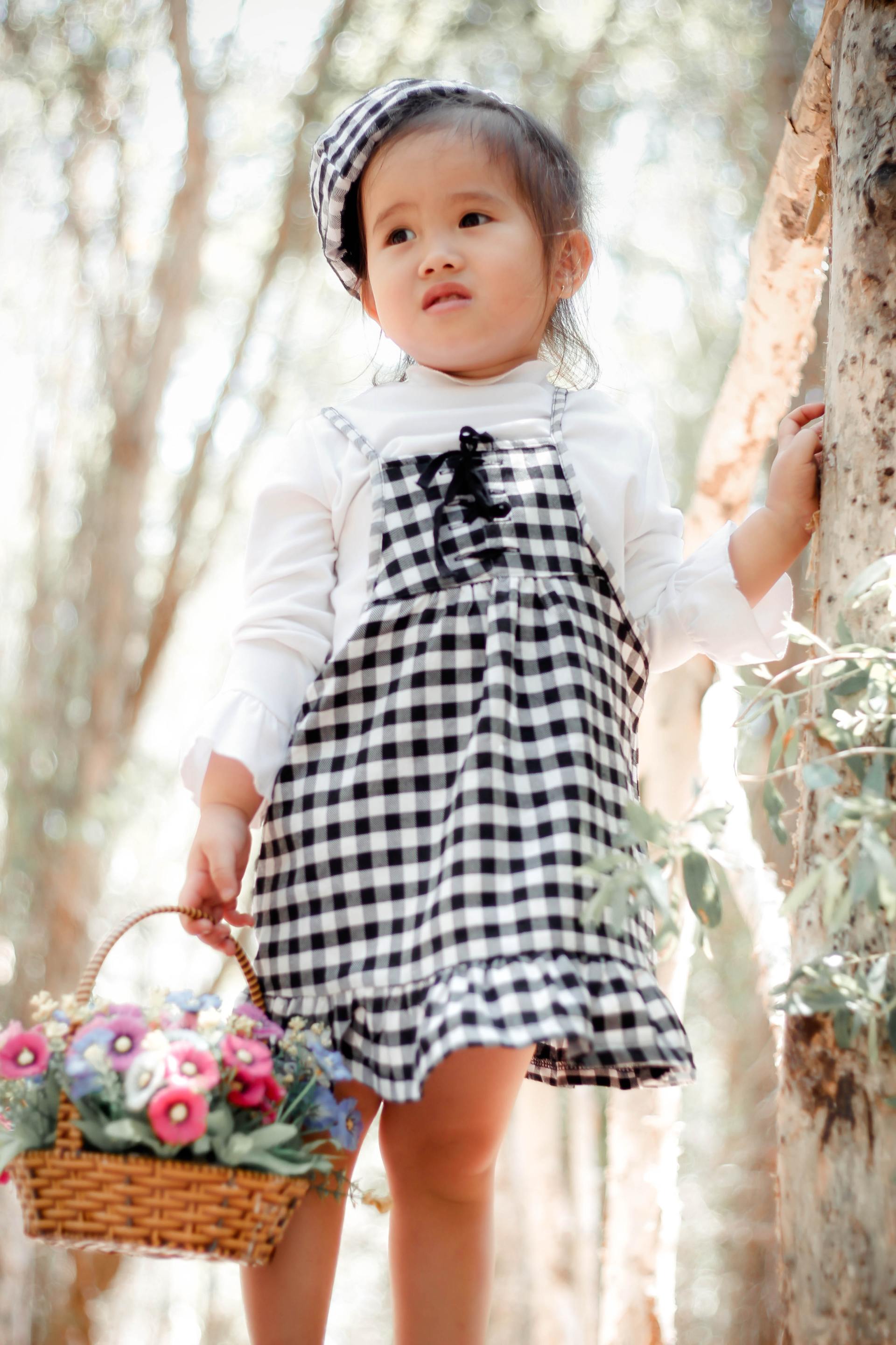 Child in a black-and-white gingham dress holding a flower basket outdoors among trees