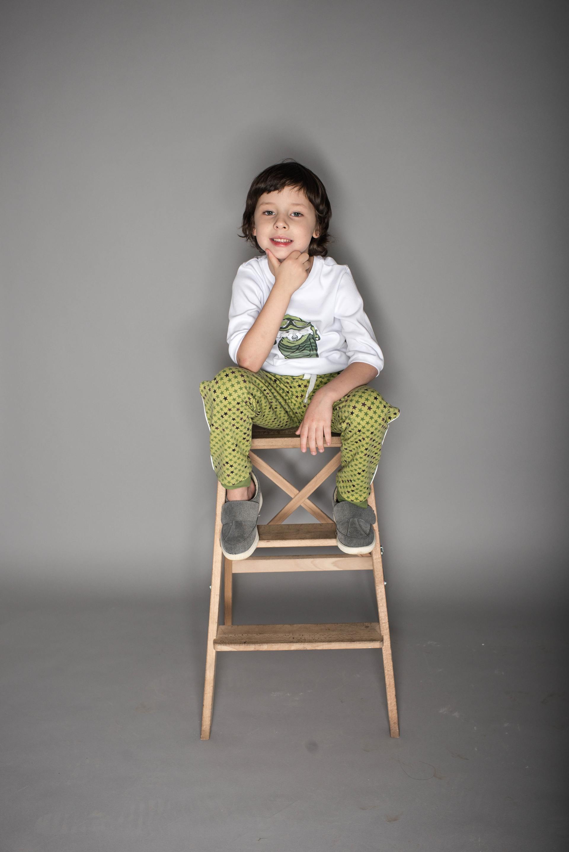 Child sitting on a wooden stool against a gray studio backdrop, smiling with hand near chin.