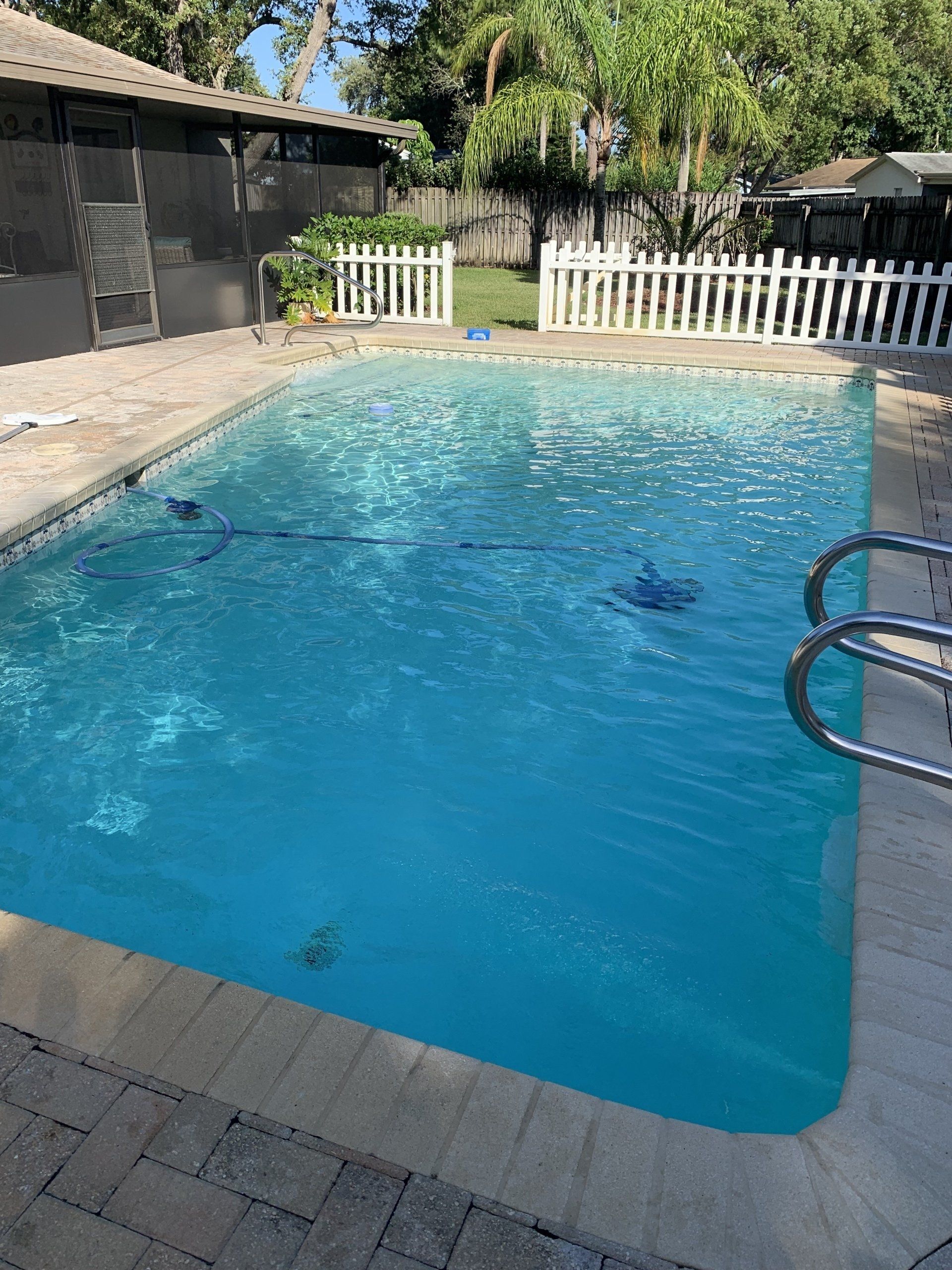Backyard swimming pool with clear blue water, white fence, and stone patio on a sunny day