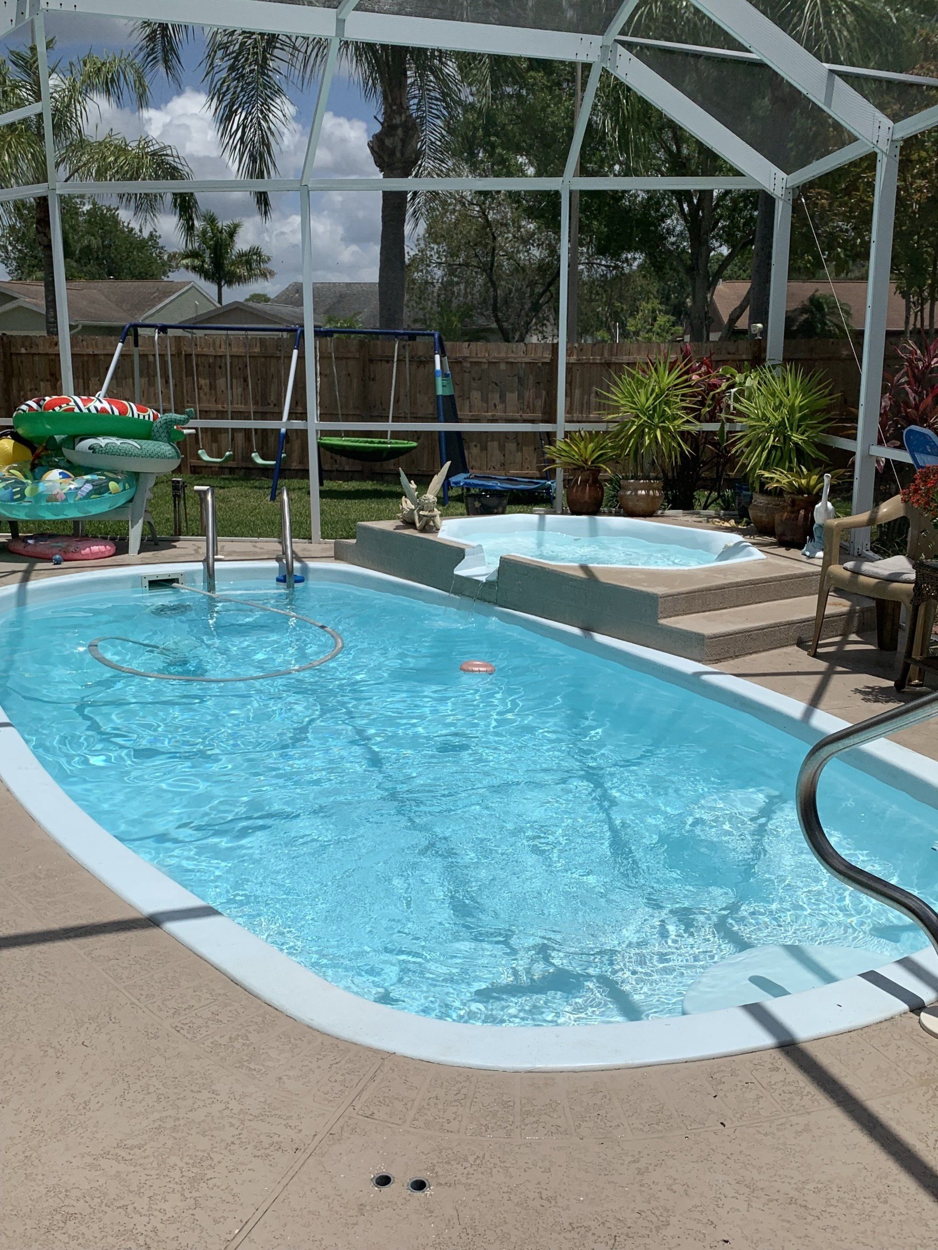 Backyard pool with clear blue water, hot tub, patio chairs, and screened enclosure under a sunny sky