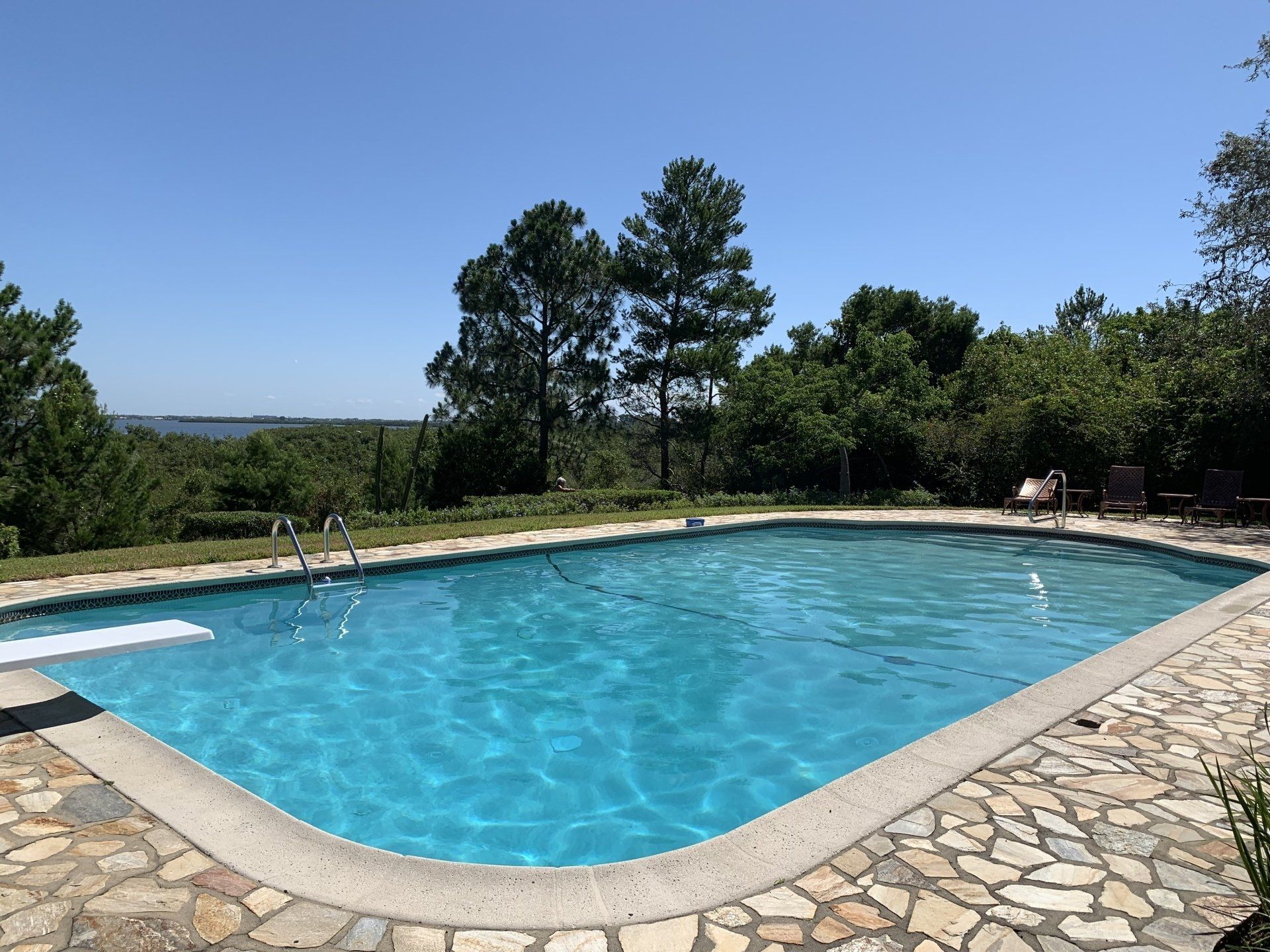 Outdoor swimming pool with clear blue water, stone deck, and trees under a sunny sky