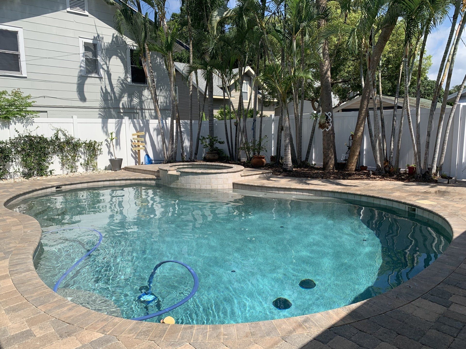 Backyard swimming pool with blue water, stone deck, and trees along a white fence