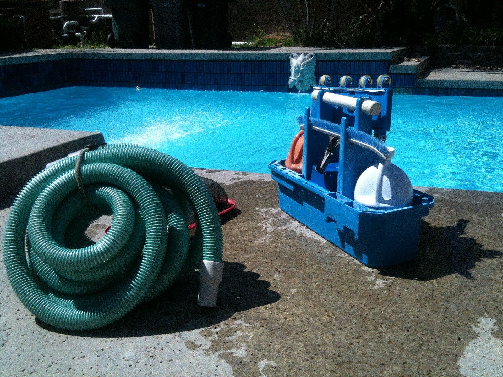 A coiled green pool vacuum hose and a blue plastic pool cleaner caddy sitting on a concrete deck beside a swimming pool.
