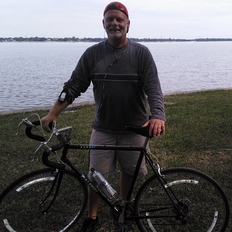 A person wearing a hat and athletic gear stands by a road bicycle on the grassy shore of a large body of water.