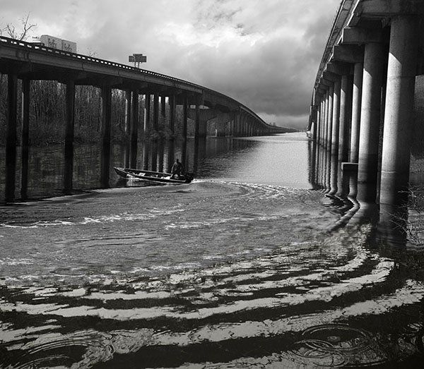 a composite image of the Atchafalaya Basin Bridge, a man in a boat and wakes in the water. Created with slight computer manipulation.
