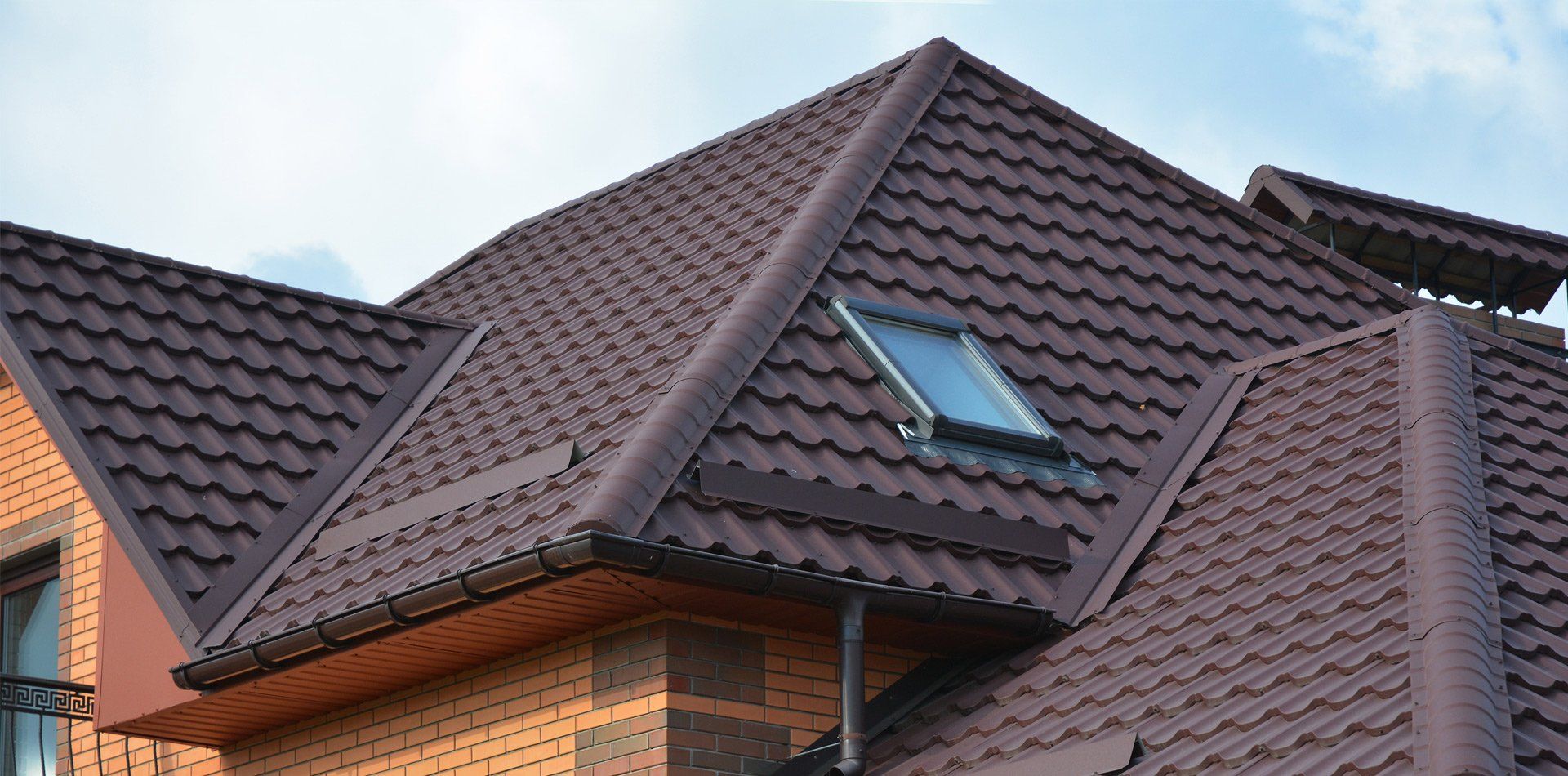 Brown tiled roof of a brick building with a skylight and a chimney against a light blue sky.